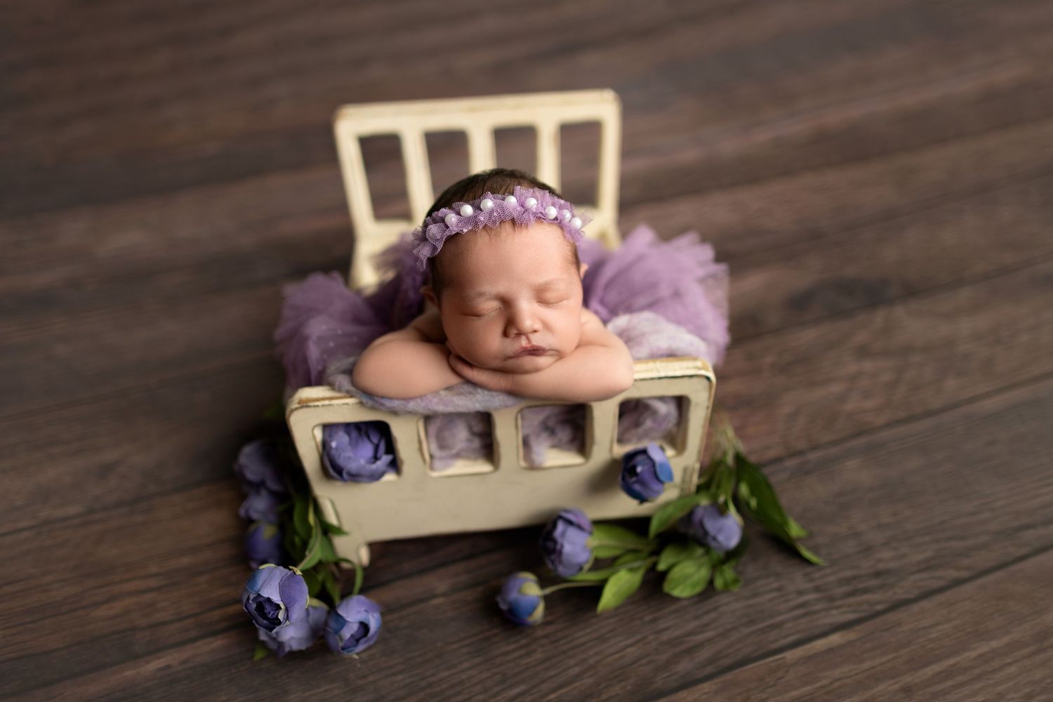 Sleeping newborn in purple outfit and bonnet, nestled in small chair with flowers.