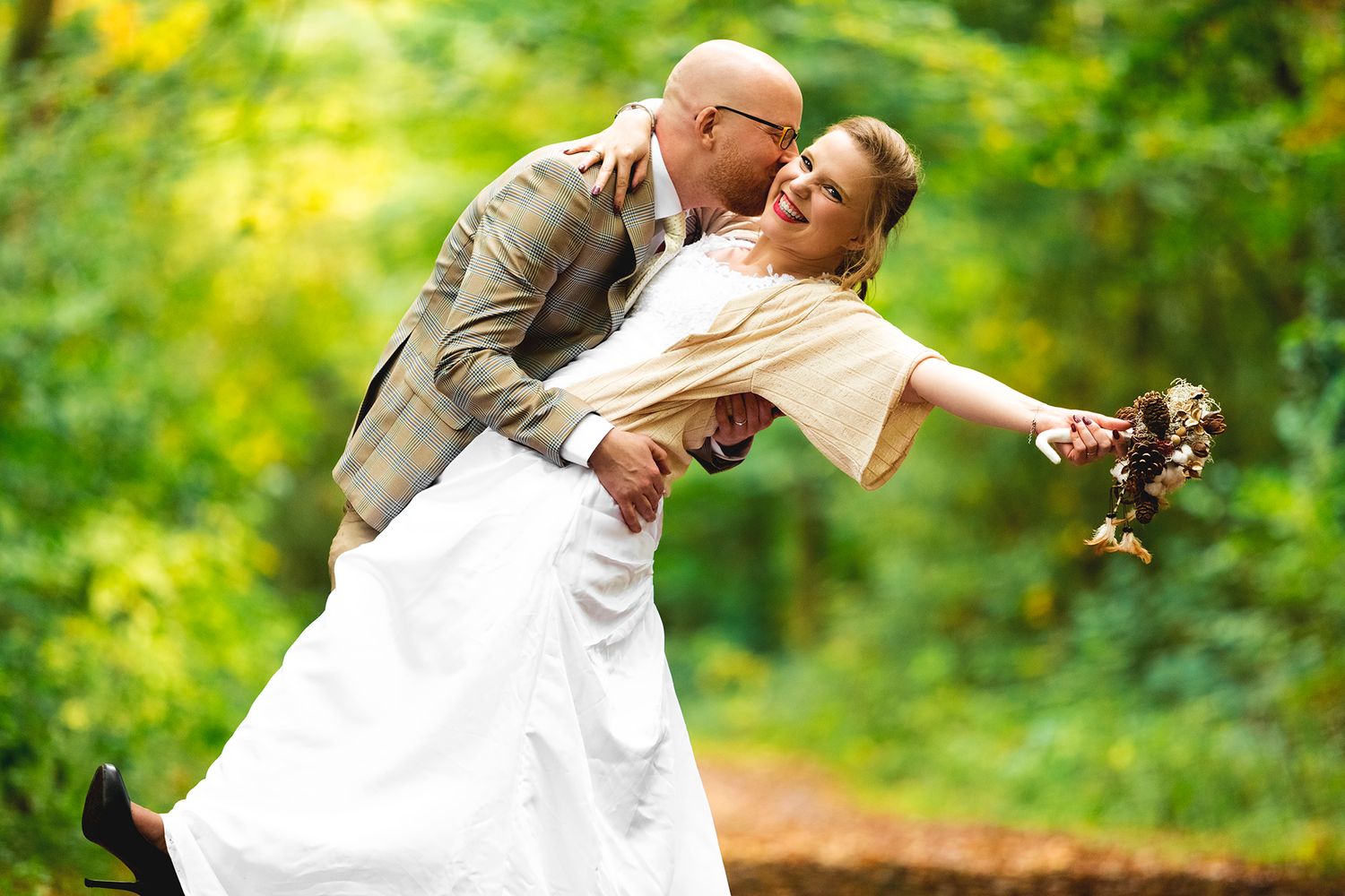 A romantic wedding couple dips in a playful pose surrounded by lush greenery outdoors.