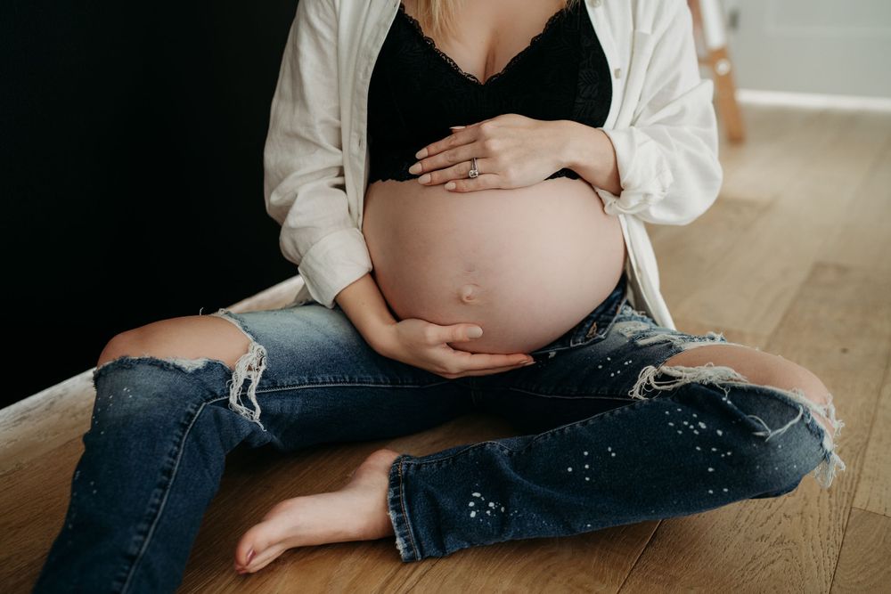 Lifestyle maternity portrait in casual denim and white cardigan on wooden floor.