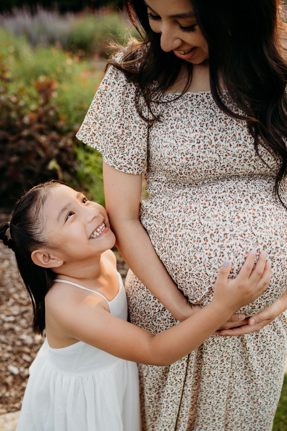 A young girl in white hugging her mother's pregnant belly in a garden setting.