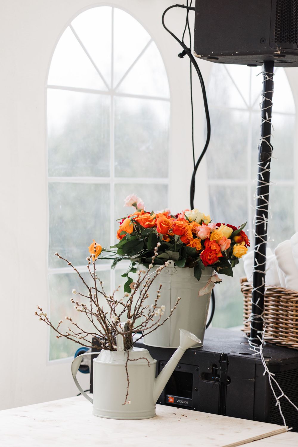 Orange flowers in white watering can by arched window with decorative lighting.