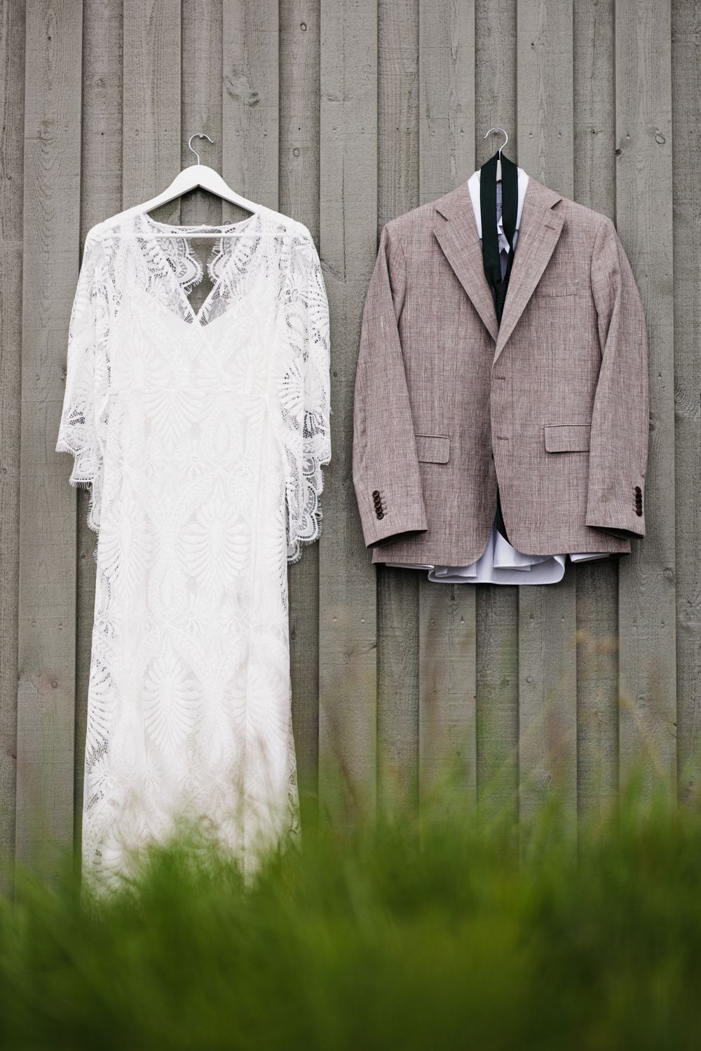 Wedding dress and gray suit jacket hanging on wooden fence outdoors.