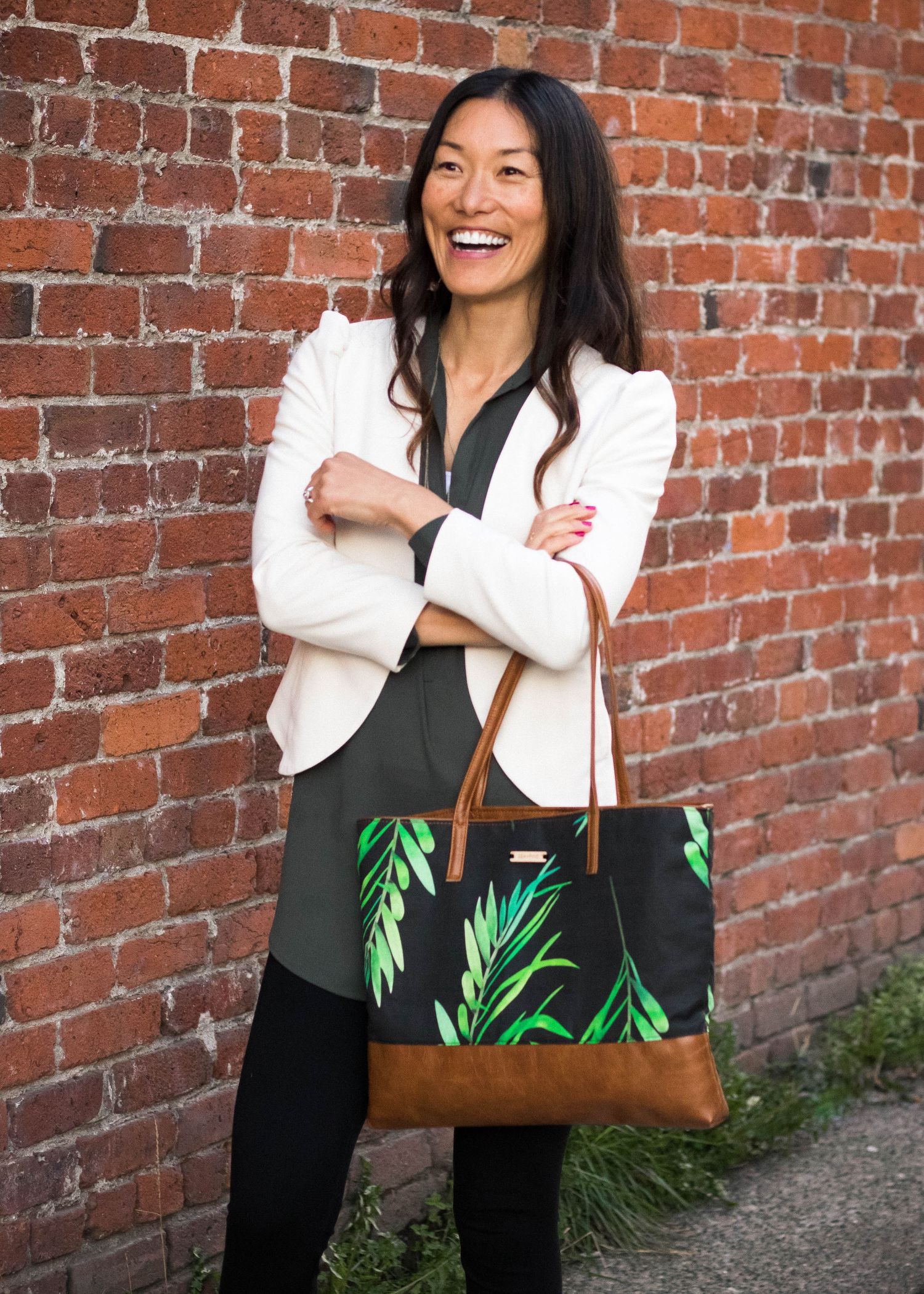 Professional woman in white blazer laughing against brick wall while holding tropical print bag.