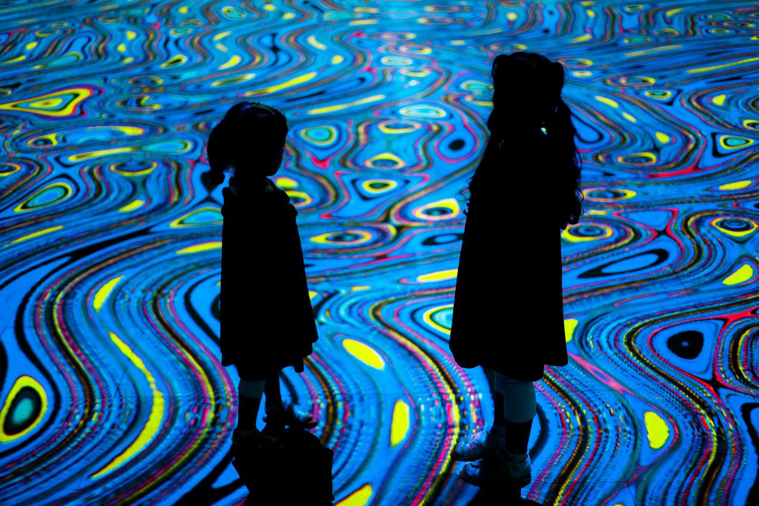 Silhouettes stand against a mesmerizing blue swirling light installation with psychedelic patterns.