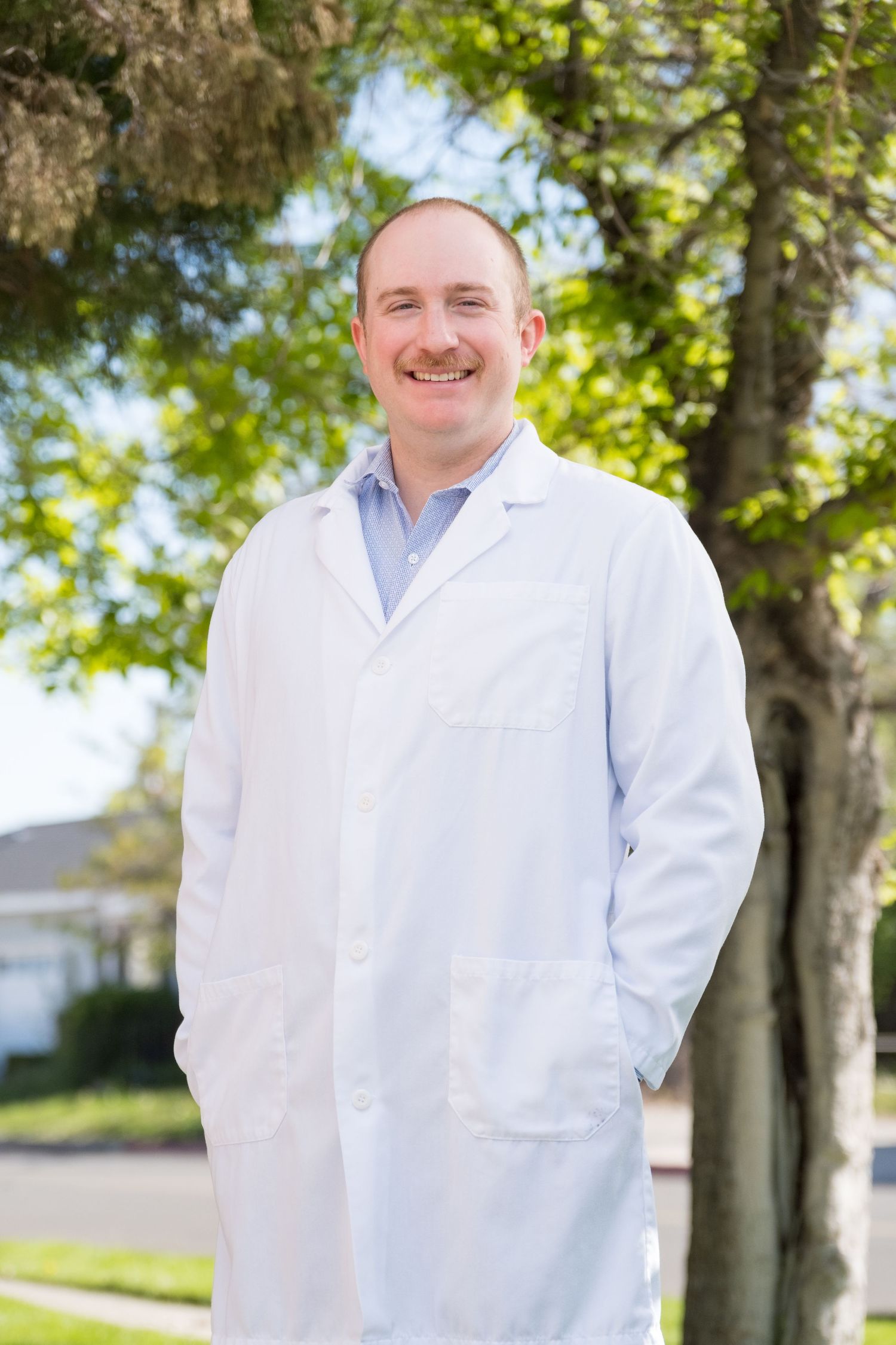 A medical professional in a white lab coat stands outdoors on a sunny day with trees in the background.
