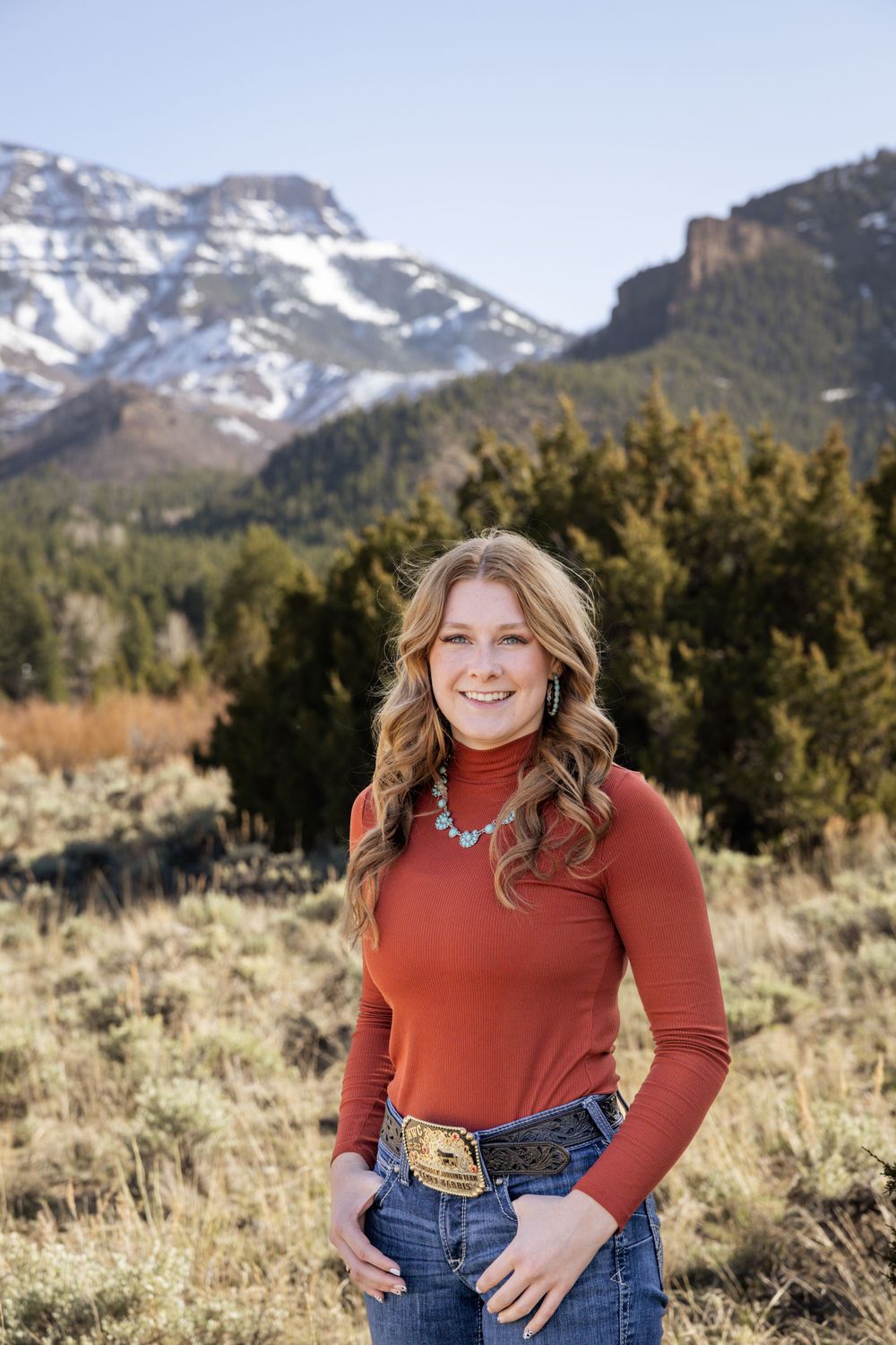A person in a red turtleneck and jeans poses against a mountain backdrop with snow-capped peaks.