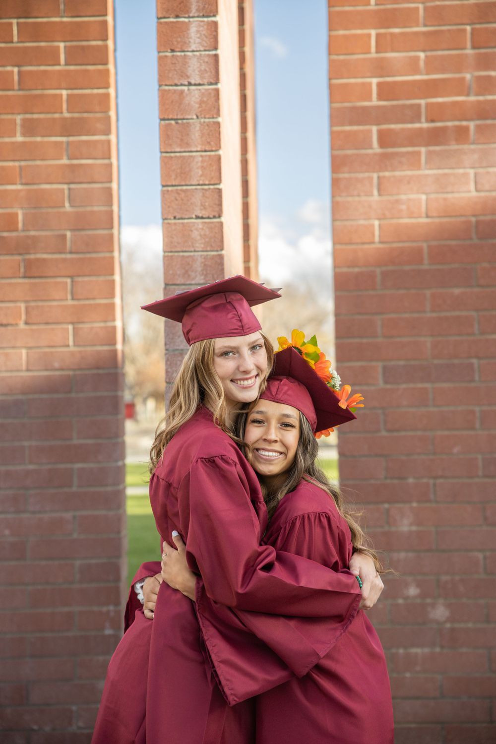 Two graduates in burgundy robes embrace while smiling against brick wall.