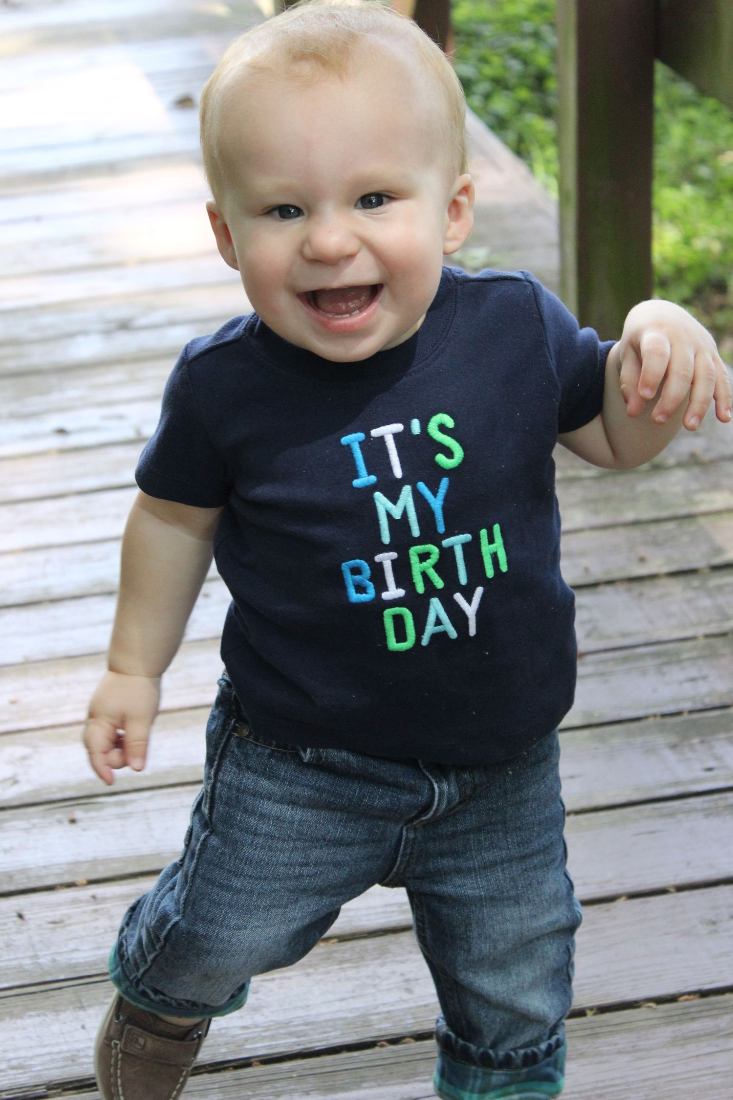 Toddler in 'It's My Birthday' shirt smiles while standing on wooden deck.