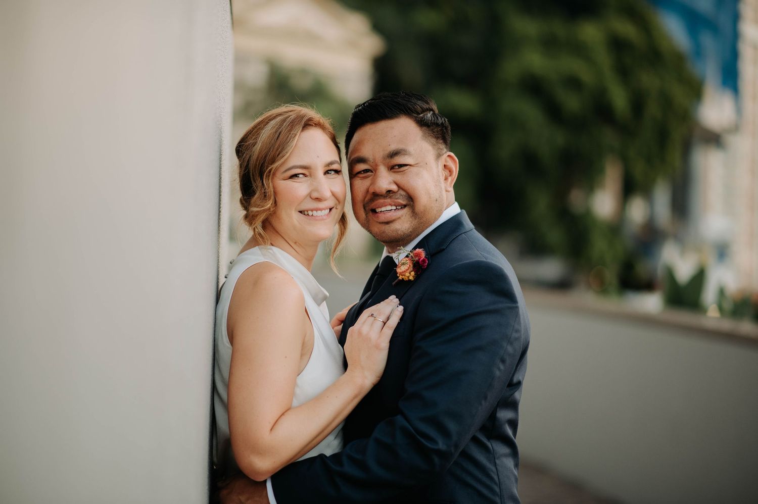 A happy couple poses together against a white wall during their wedding day.