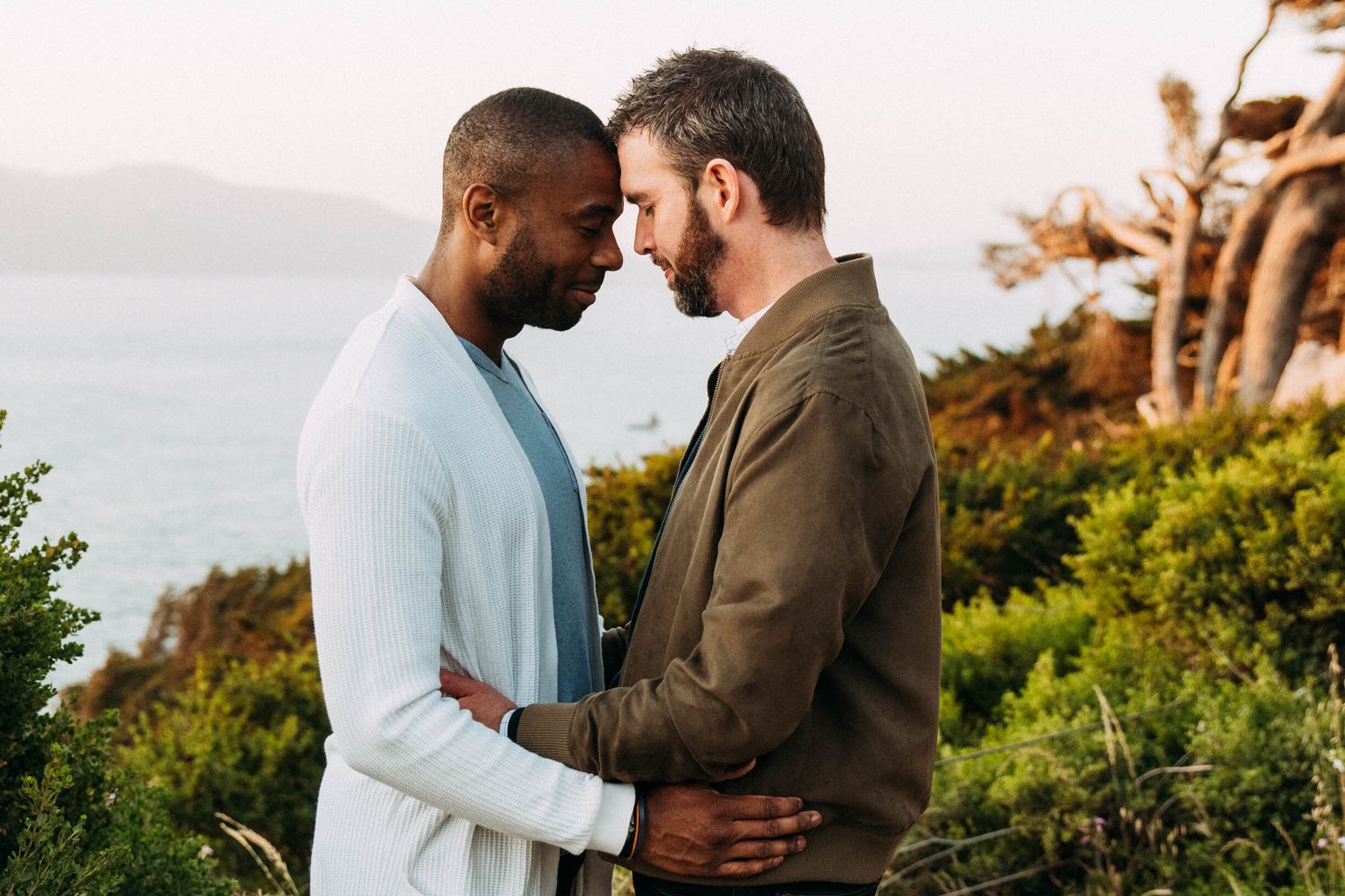 Gay, LGBTQ+, and bi-racial couple lean heads towards each other along Lands End in San Francisco for engagement photos.