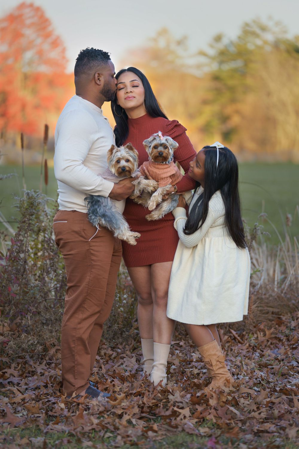 Couple in casual fall outfits poses with their two small dogs among autumn leaves.