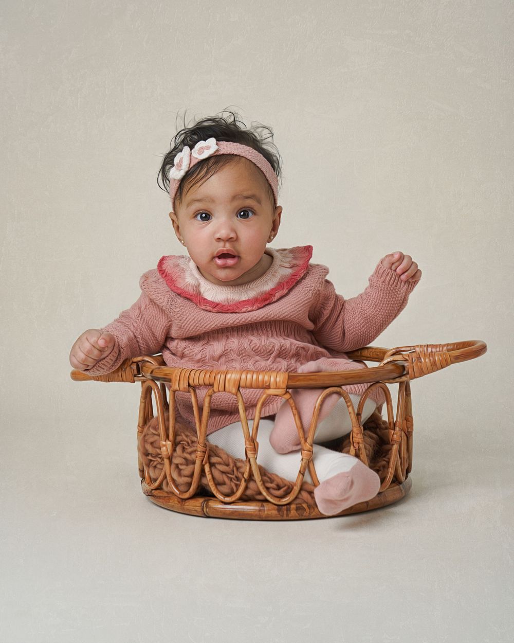 Baby in pink dress sitting in woven rattan basket during studio photoshoot.