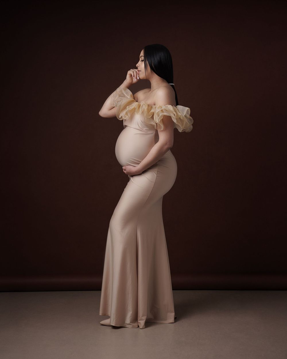 Maternity portrait in nude dress with ruffled shoulders against dark background.