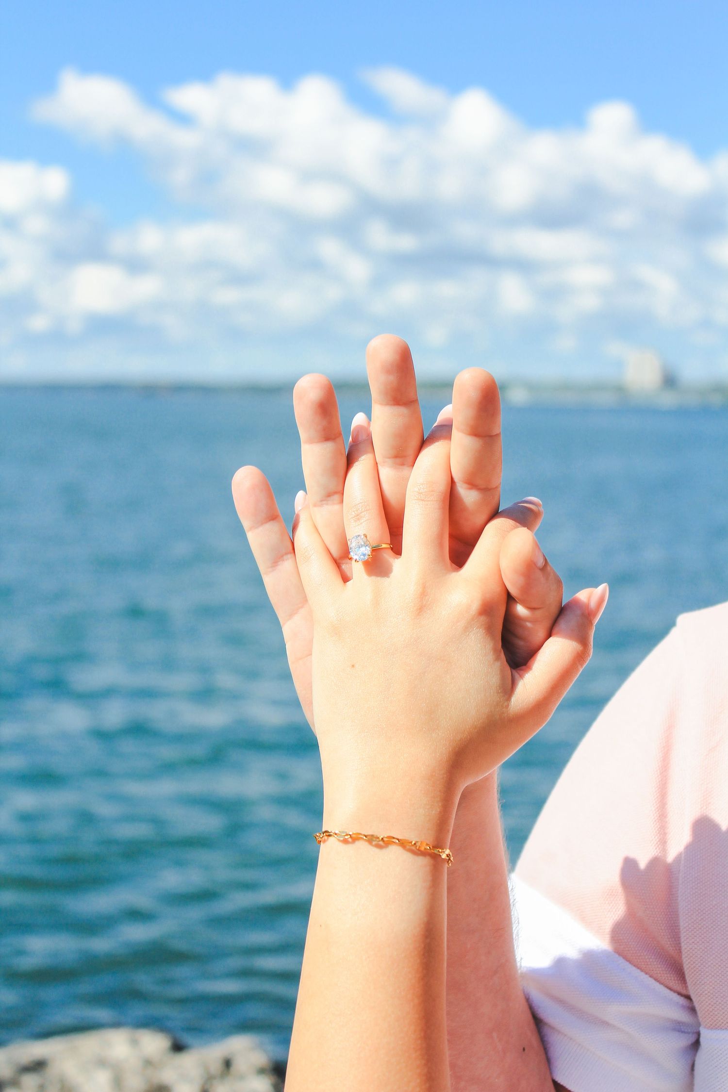 Two hands intertwined showing off an engagement ring against a beautiful ocean backdrop.