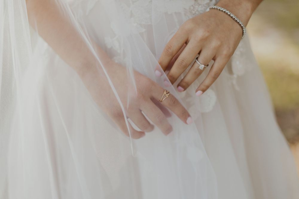 Close-up detail of wedding rings and jewelry on hands against white dress fabric.