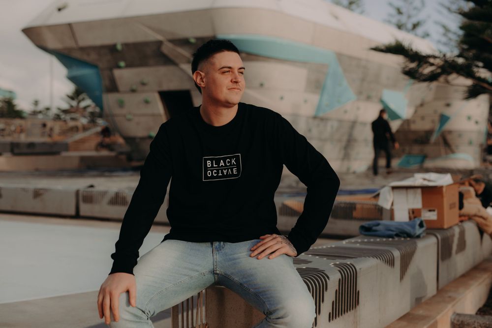 A person in a black sweater sits casually on a concrete wall near the ocean.