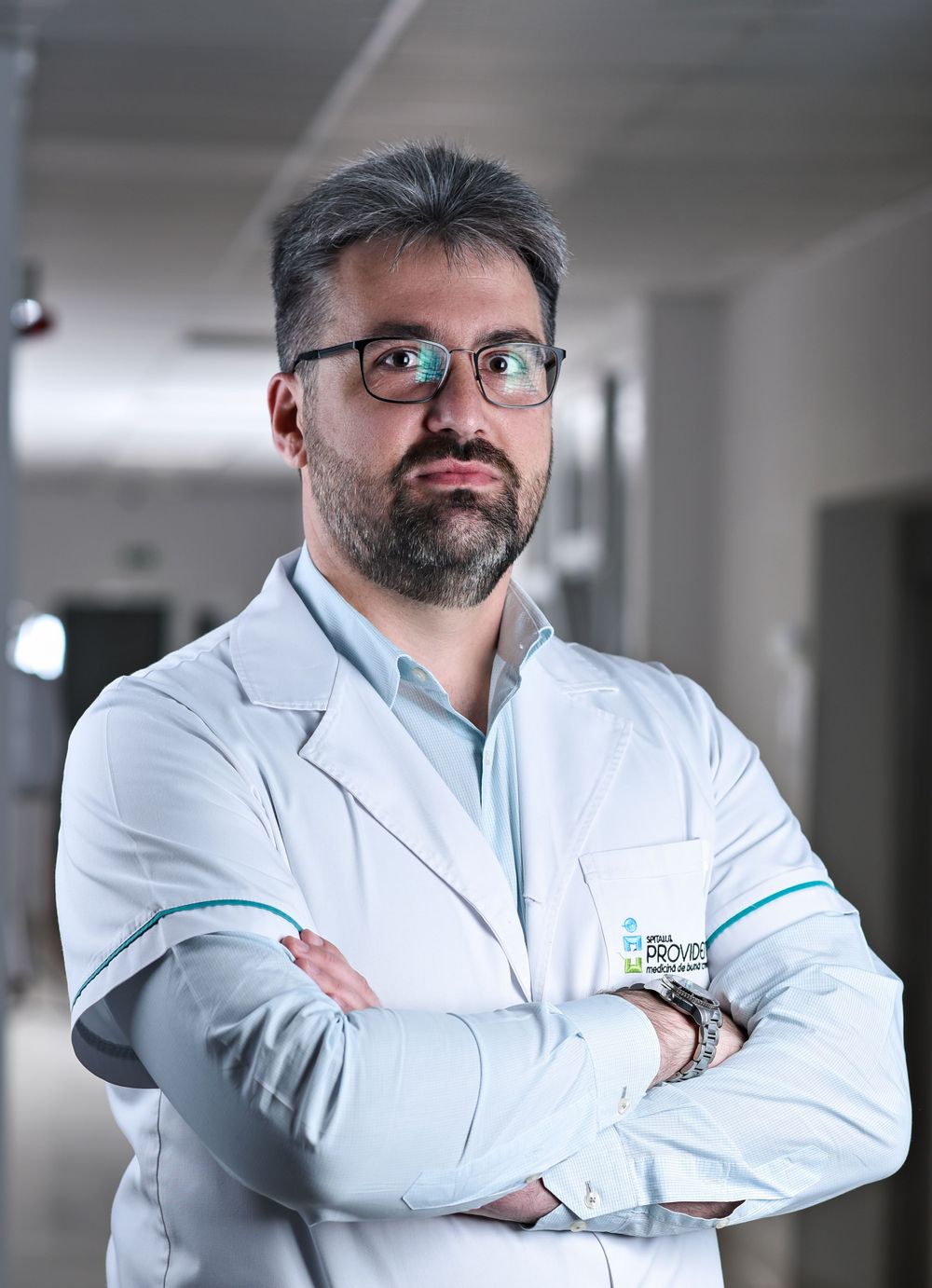 Medical professional in white coat and glasses standing in hospital hallway.