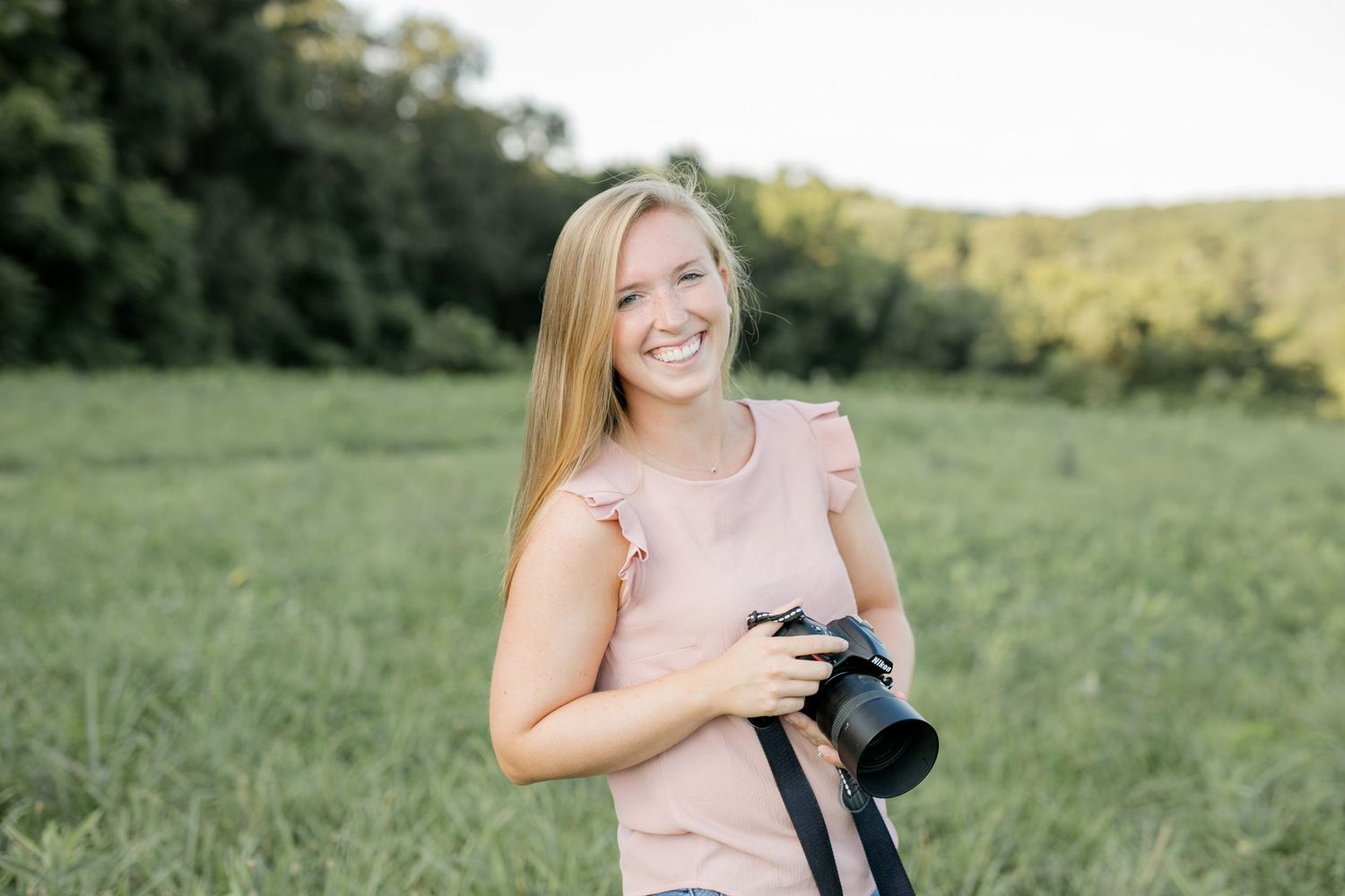 A photographer stands in a sunlit meadow holding professional camera equipment.