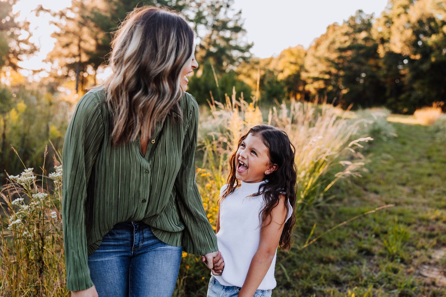 A mother and young daughter laugh together in a sunlit field surrounded by tall grasses and wildflowers.