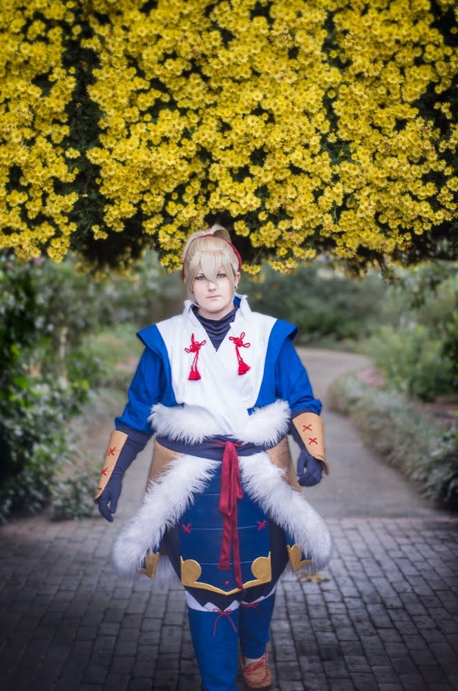 A cosplayer in a blue and white costume standing in front of blooming yellow flowers.