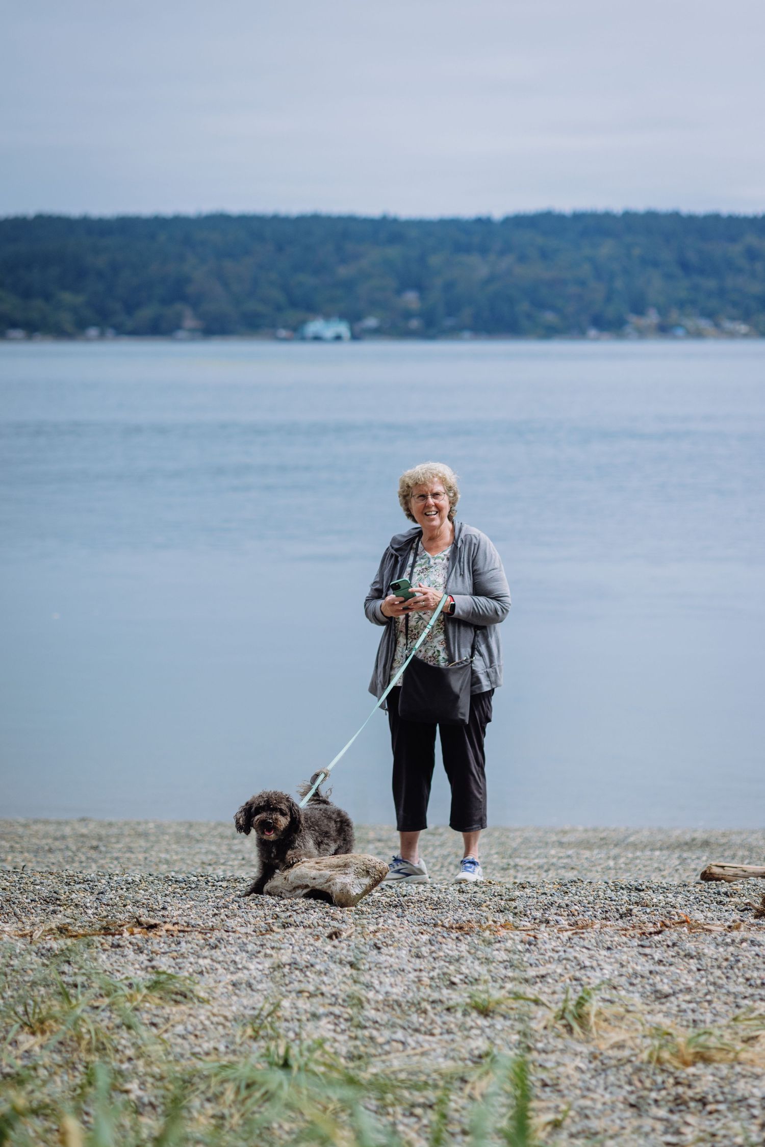 Senior woman with curly hair walks her shaggy dog on a rocky beach with calm water and hills behind her.