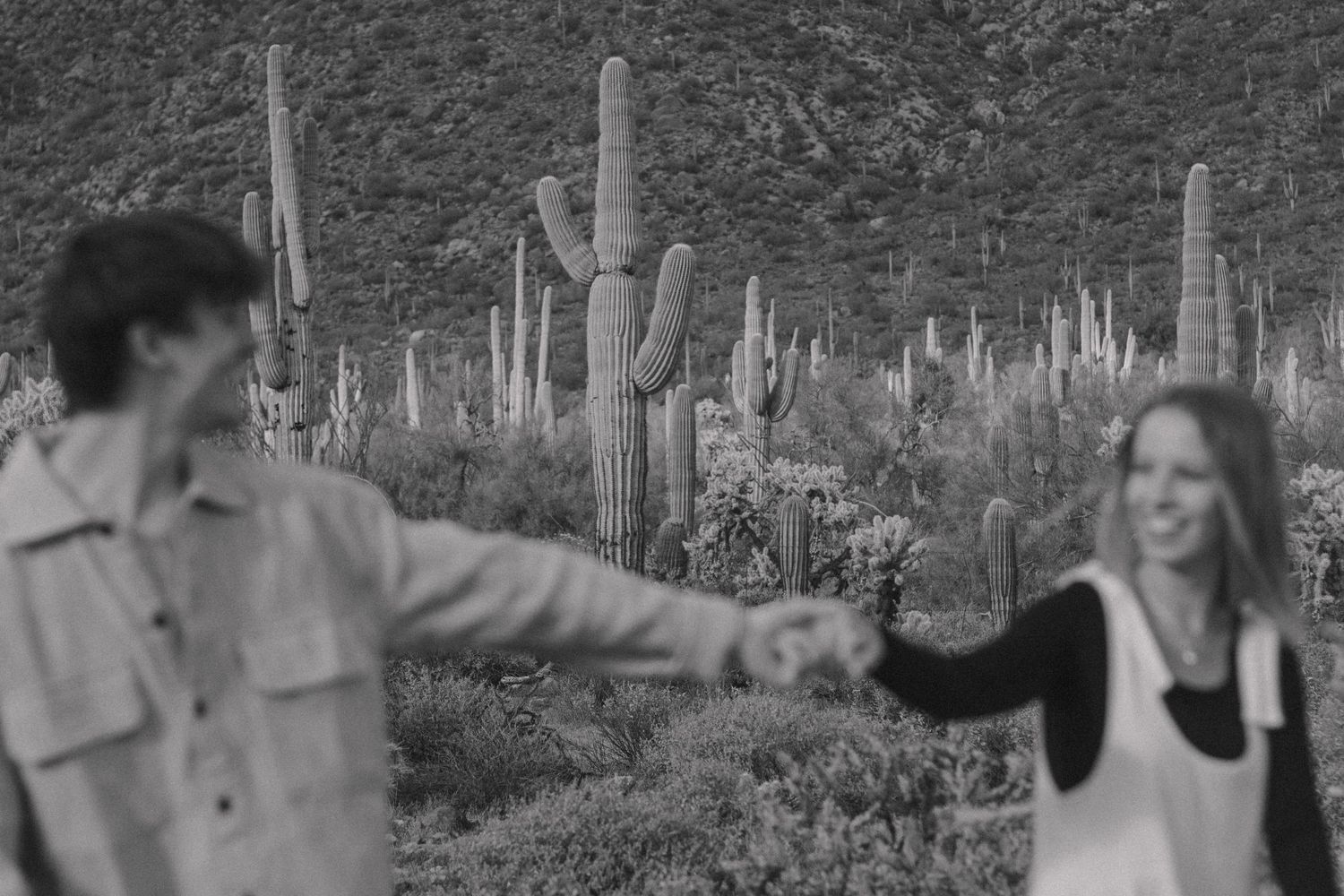 Black and white photo of two people reaching toward each other with saguaro cacti in the background.