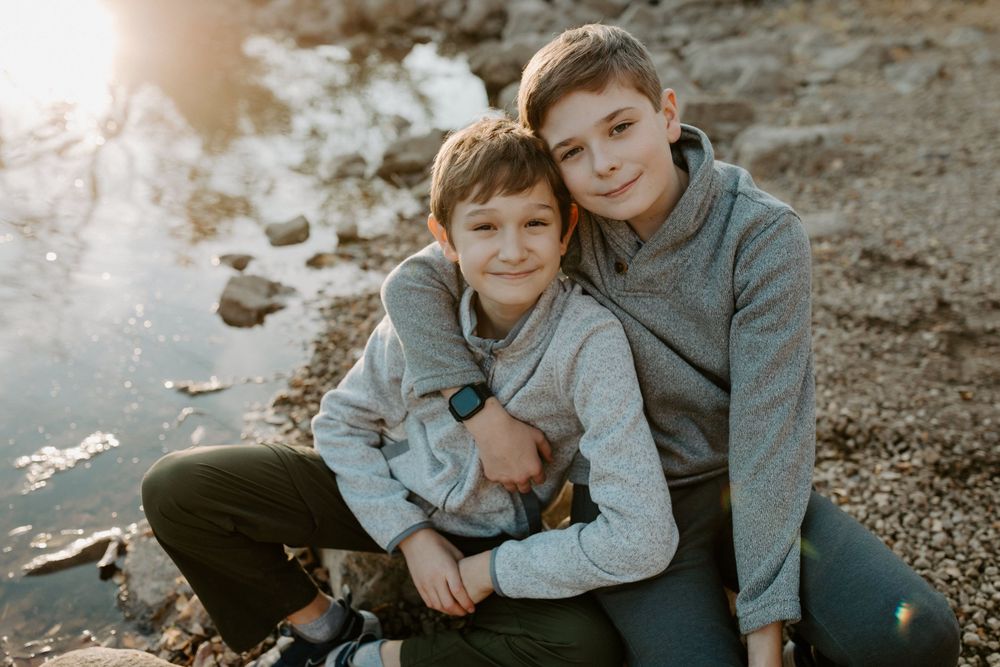 Two young boys in grey sweaters share a warm embrace by rocky shore at sunset.