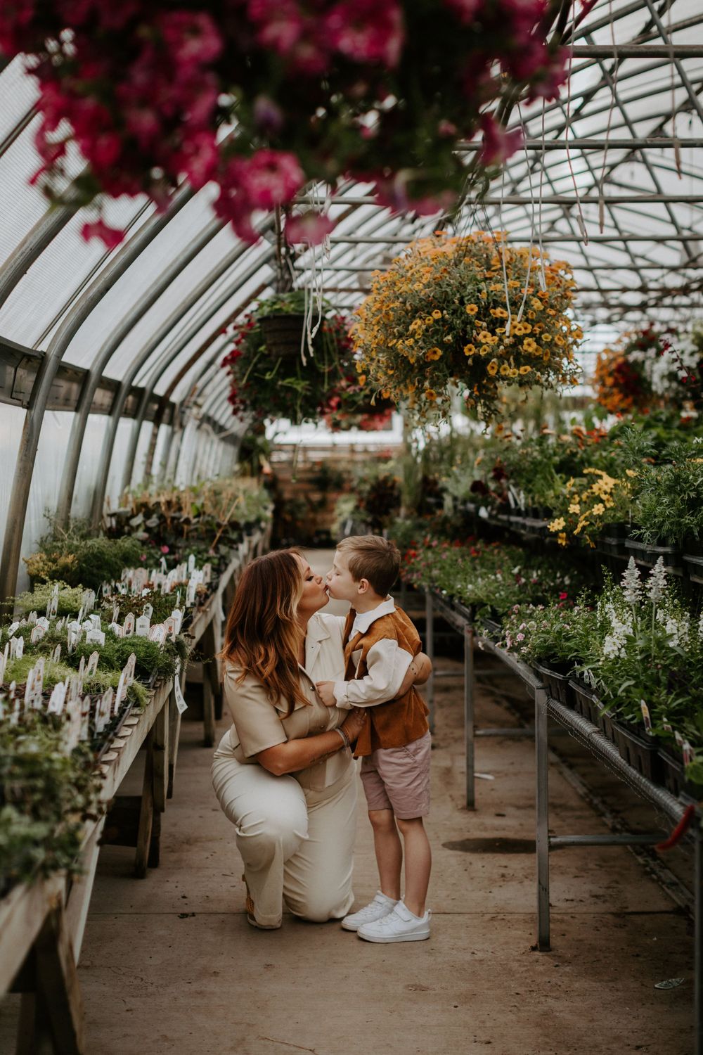 A tender family moment inside a greenhouse filled with colorful hanging flowers and blooming plants.