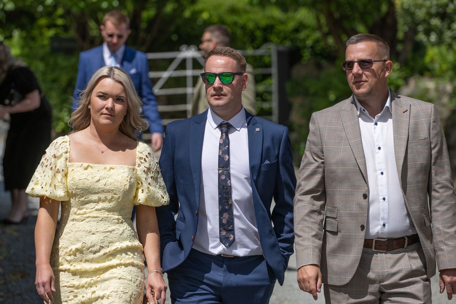Three wedding guests in formal attire walking together on a sunny outdoor path.