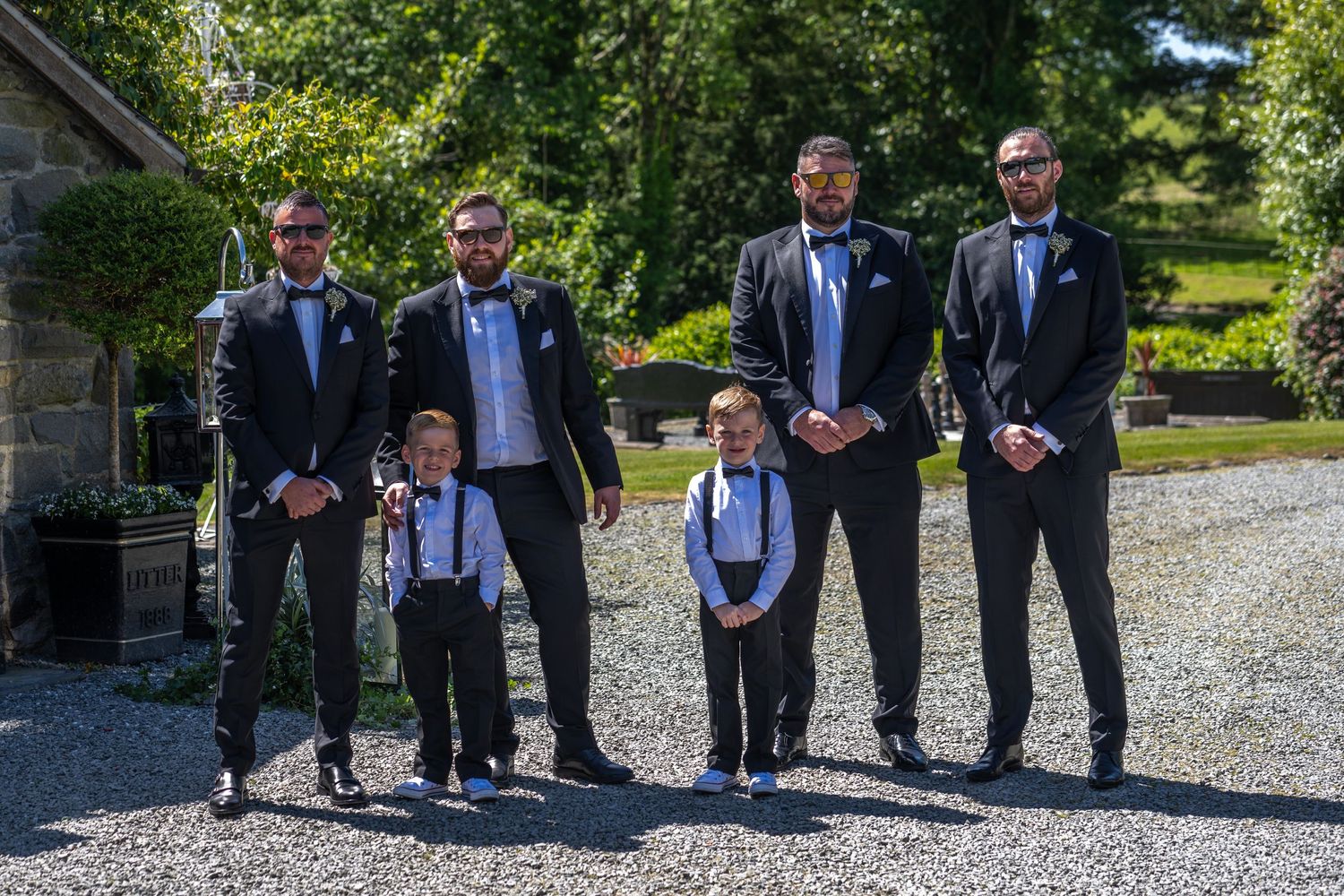 Groomsmen and young attendants in matching suits pose for a formal photo on a gravel path.