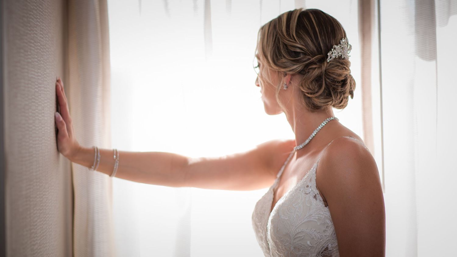 Bride wearing a lace gown and pearl accessories gazes out a sunlit window on her wedding day.