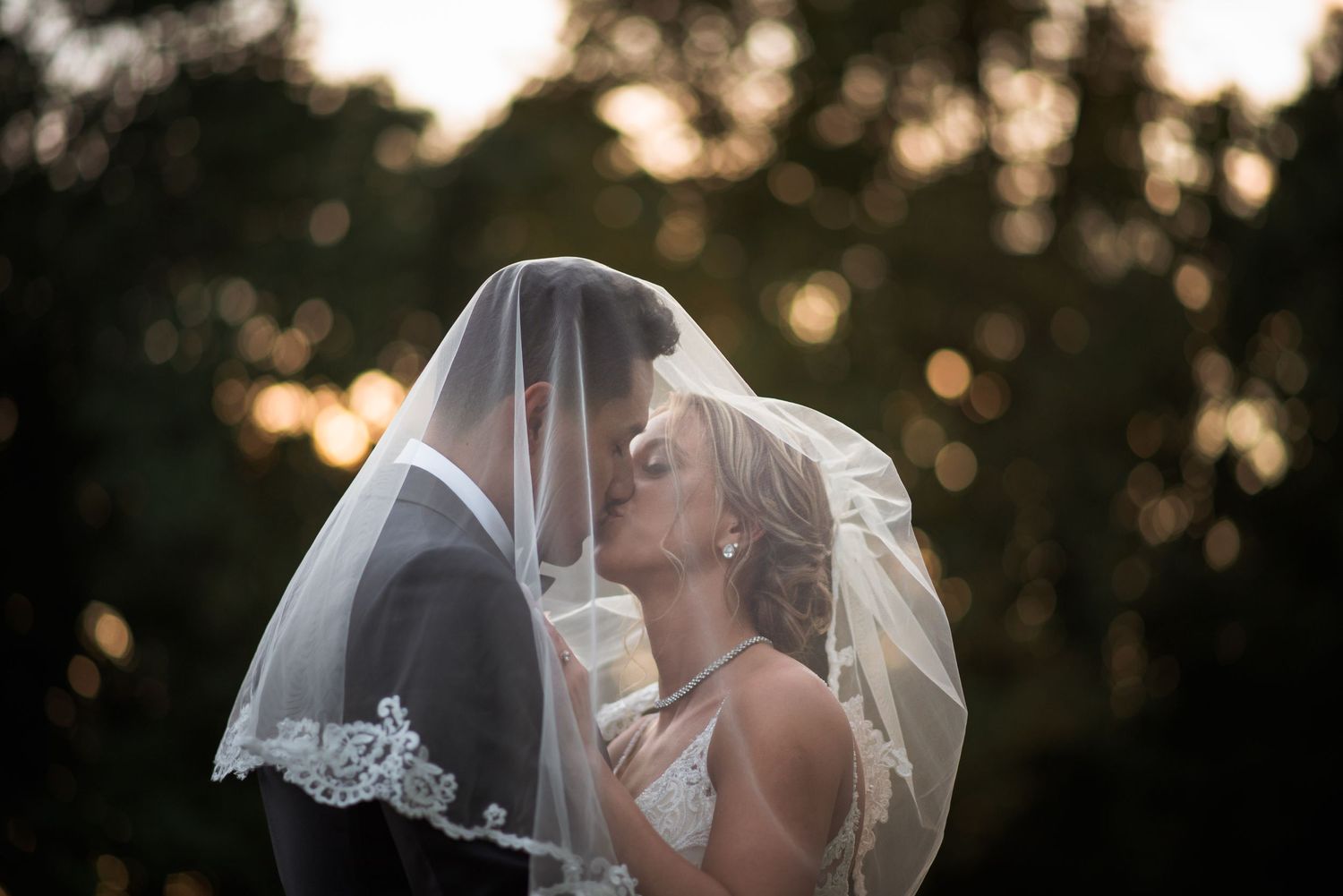 Bride and groom share intimate kiss under lace veil in golden sunset light.