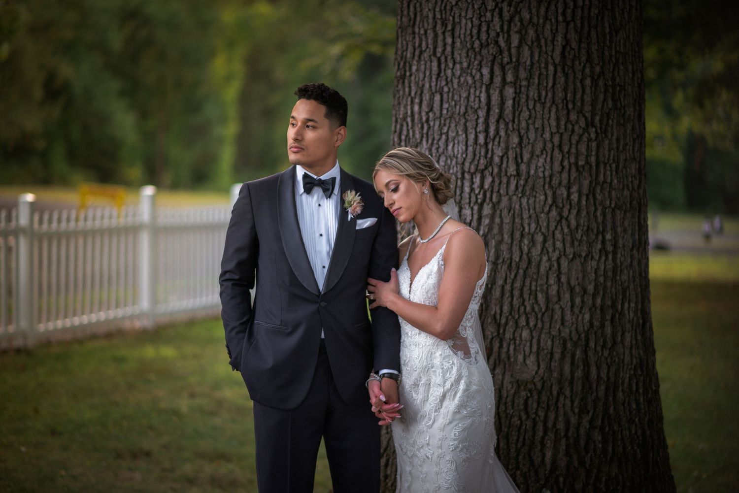Bride leans on groom in wedding attire beside large tree at outdoor venue.