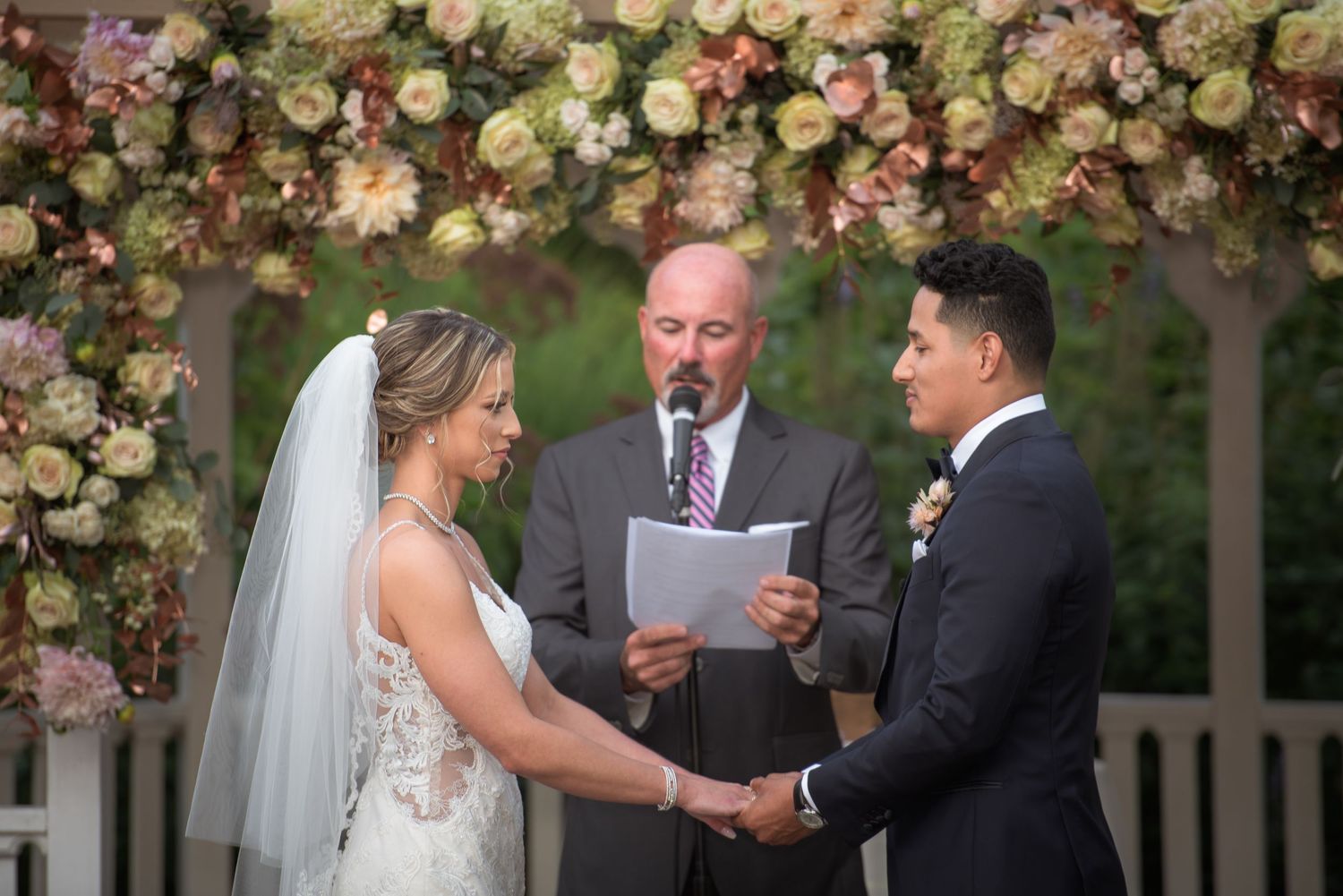 Bride and groom exchange vows at outdoor wedding ceremony beneath stunning floral arch with officiant reading from paper.