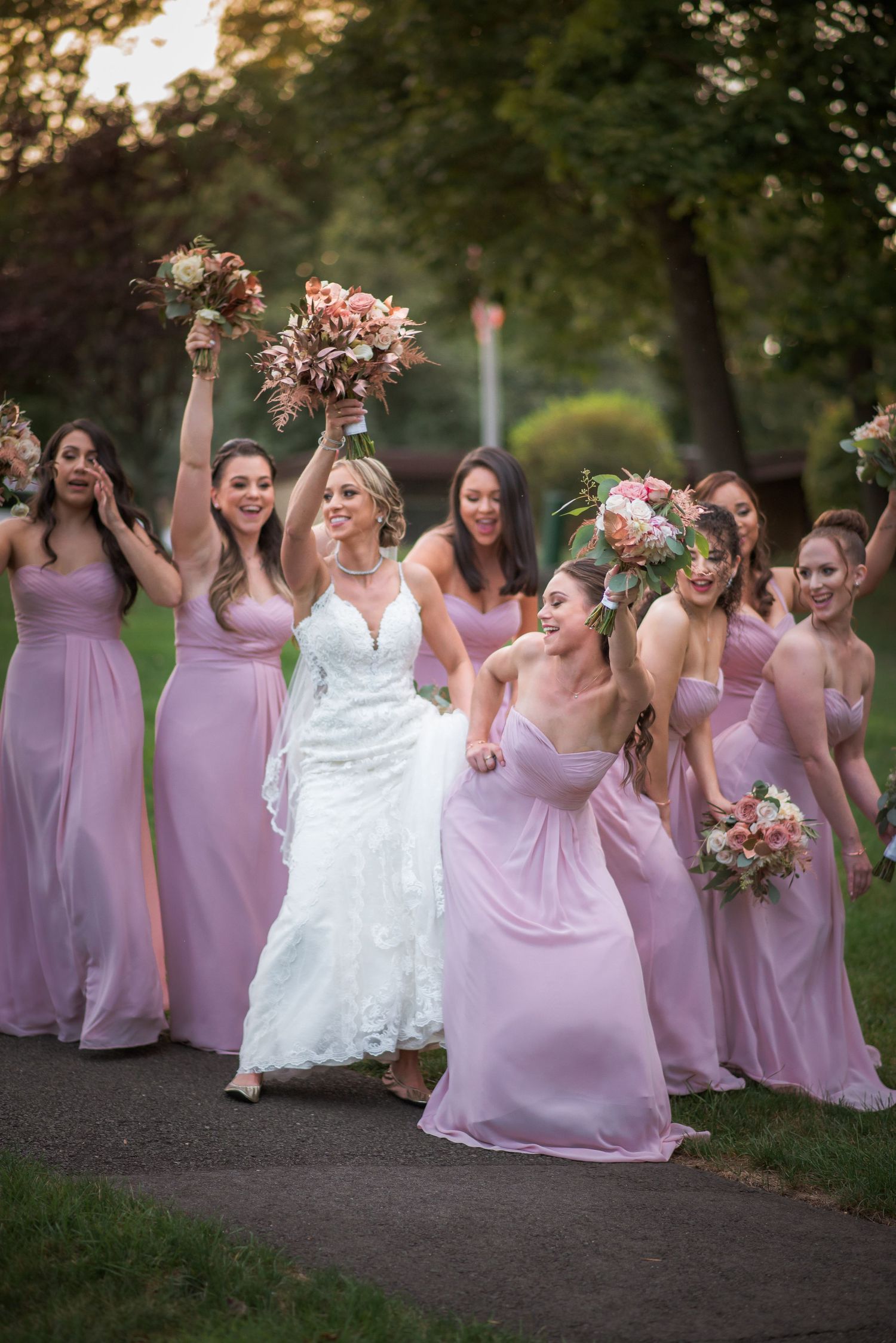 Bride celebrates with bridesmaids in pink gowns raising bouquets on garden path.