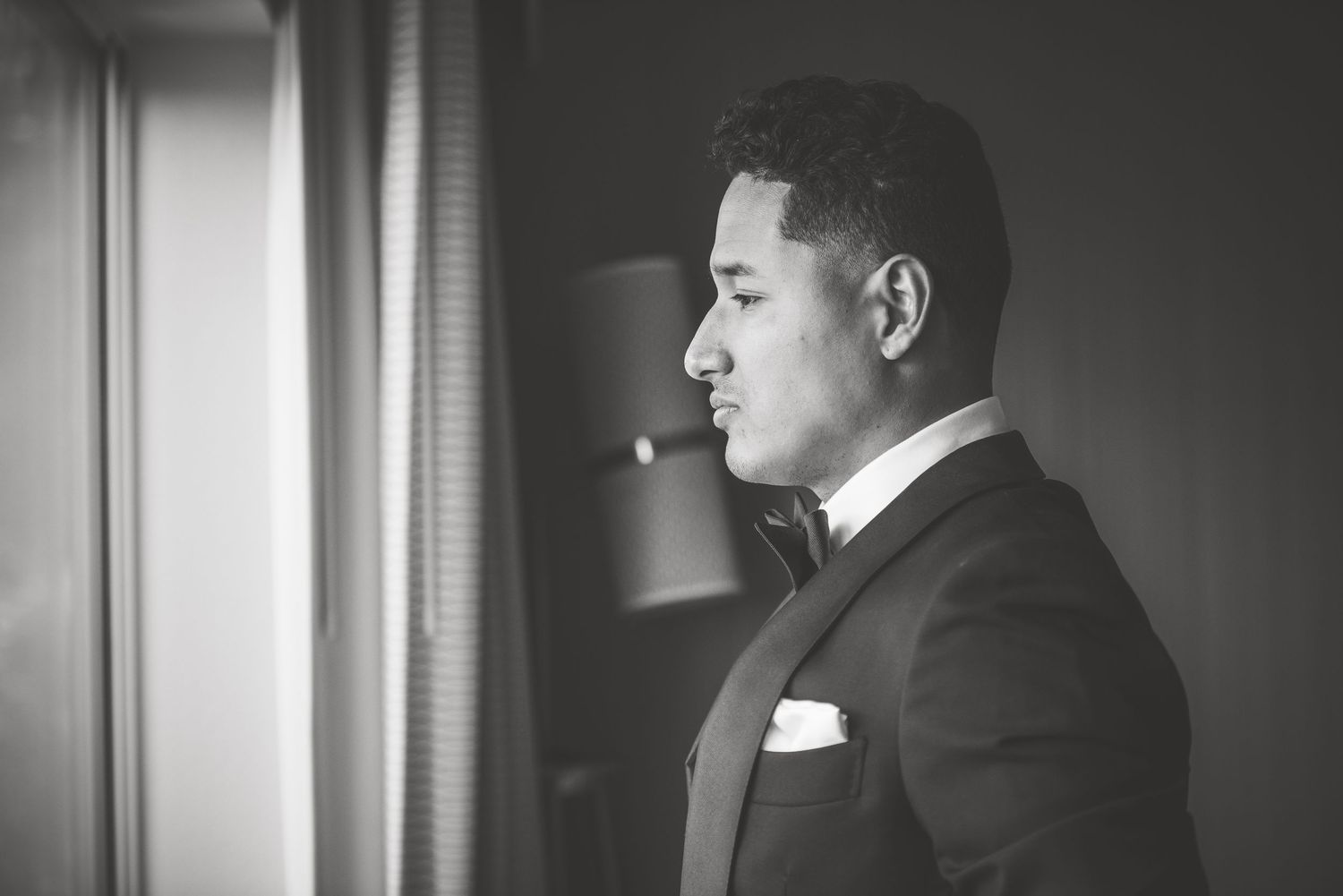 Elegant groom in black tuxedo with bow tie standing by window in moody black and white wedding portrait.
