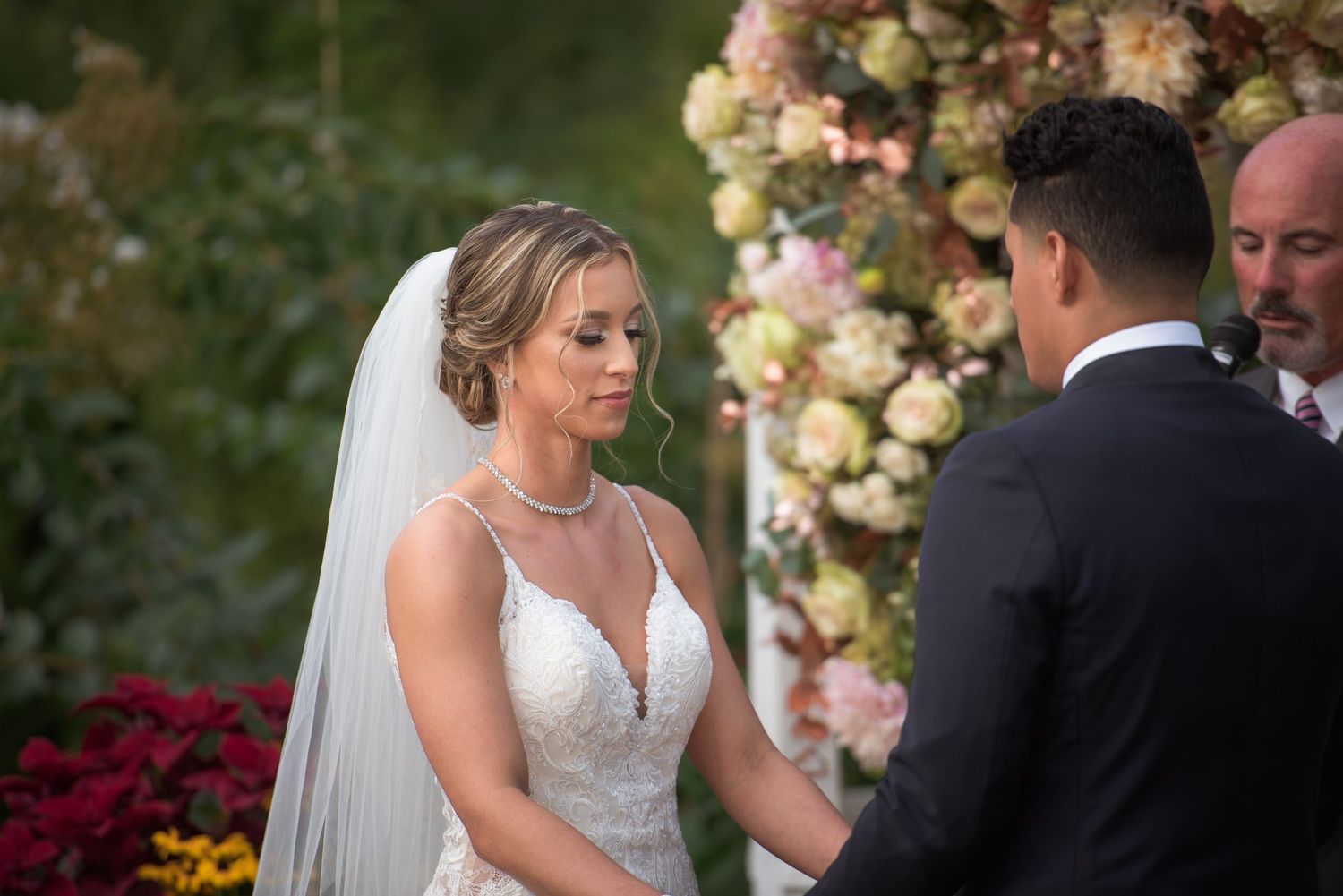 Tearful bride in lace wedding gown and veil holds grooms hands during heartfelt outdoor wedding ceremony.