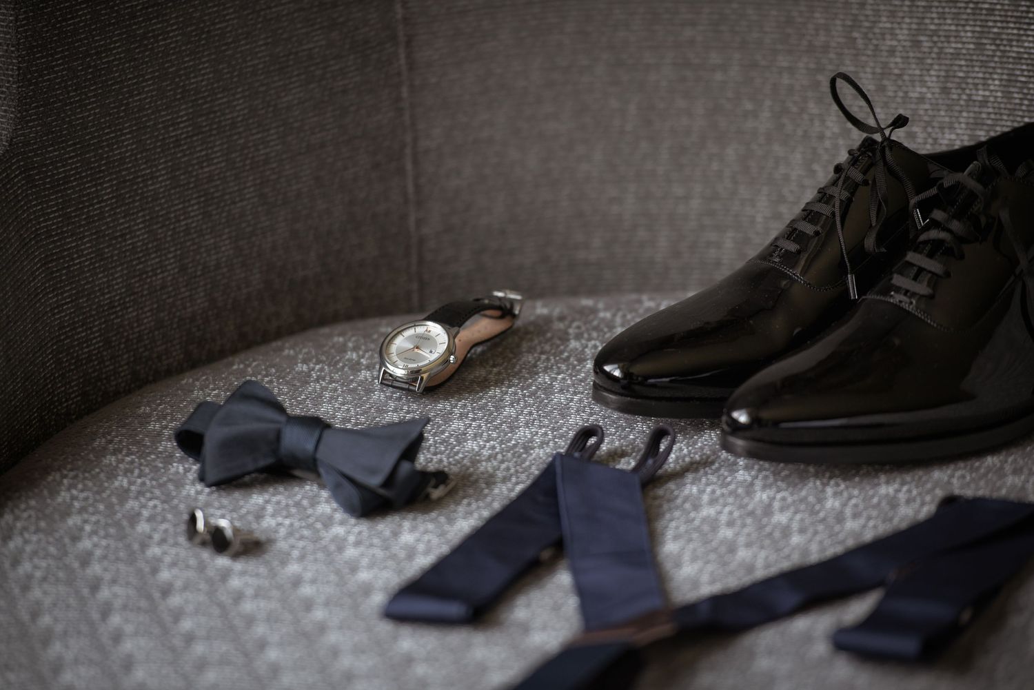 Elegant groom accessories including black patent leather dress shoes, navy bow tie, suspenders, watch, and cufflinks arranged on gray chair.