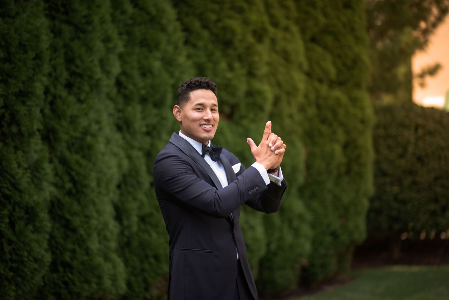 Handsome groom in navy tuxedo smiling and clapping hands outdoors with lush green hedges in background.