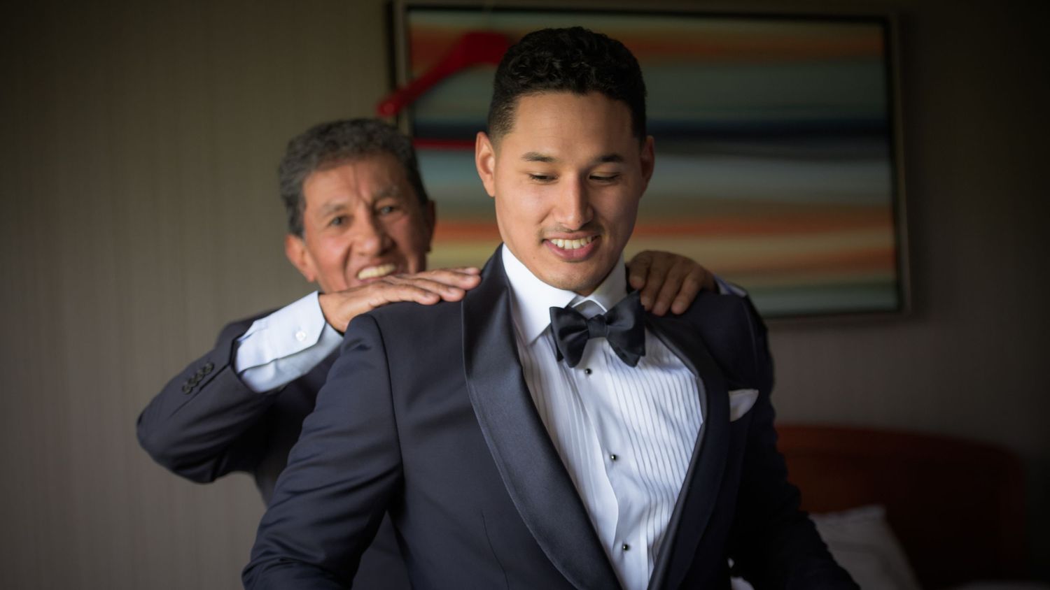 Older man helping young groom in navy tuxedo adjust his bow tie during wedding day getting ready preparations.