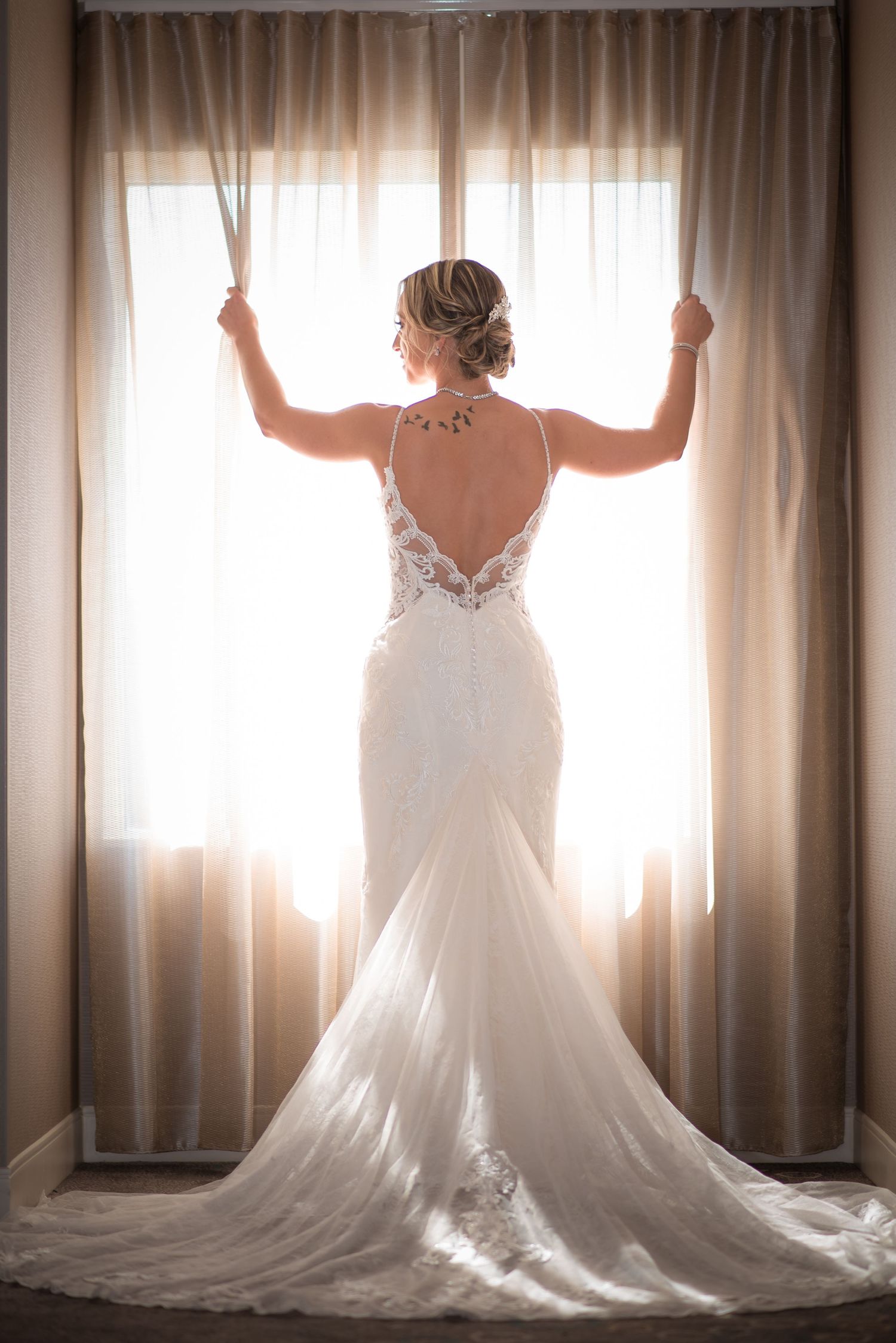 Bride in a stunning backless lace wedding gown stands before a sunlit window holding the curtains.