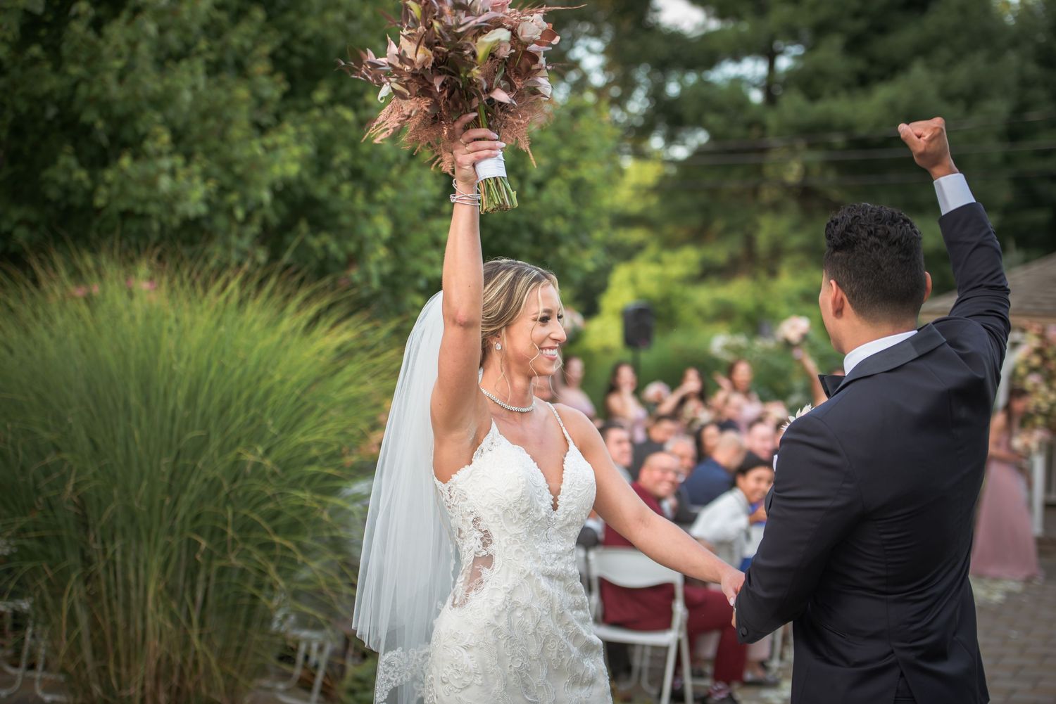 Joyful bride raises bouquet as couple celebrates after outdoor wedding ceremony.