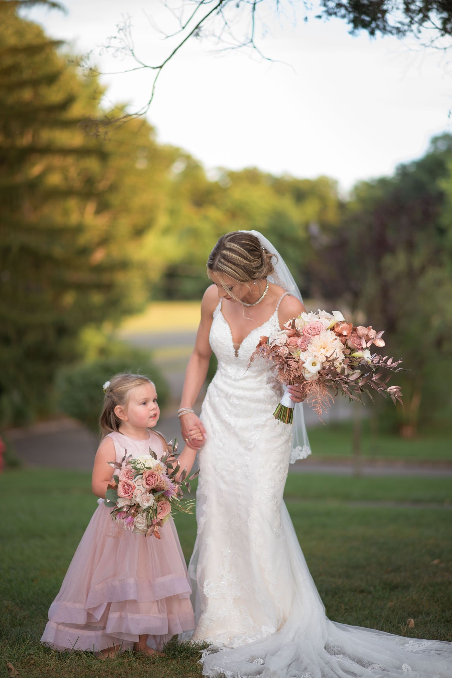 Bride in lace gown holds bouquet while posing with flower girl in pink dress.