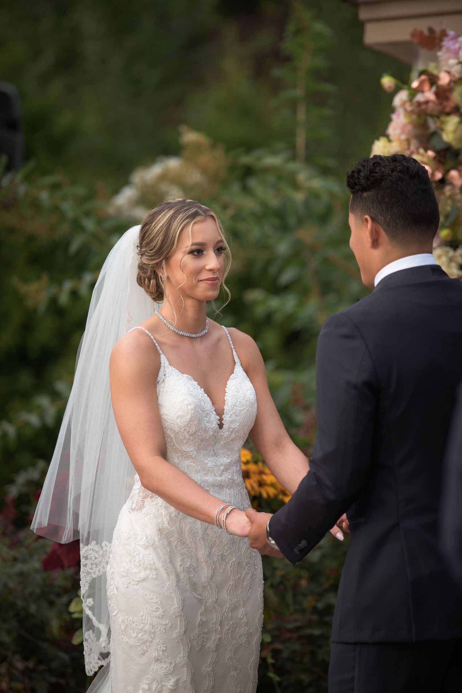 Bride in lace wedding gown and veil holds hands with groom during outdoor wedding ceremony.