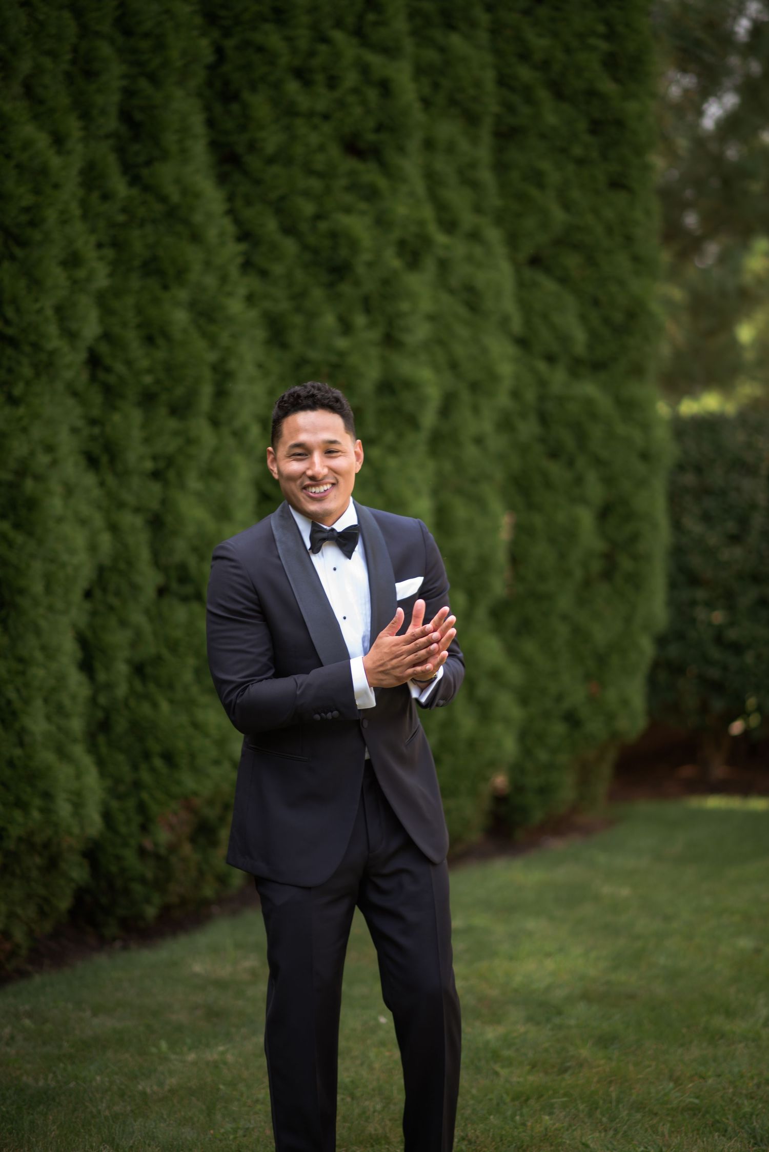 Smiling groom in navy tuxedo with bow tie and pocket square standing on green lawn before wedding ceremony.