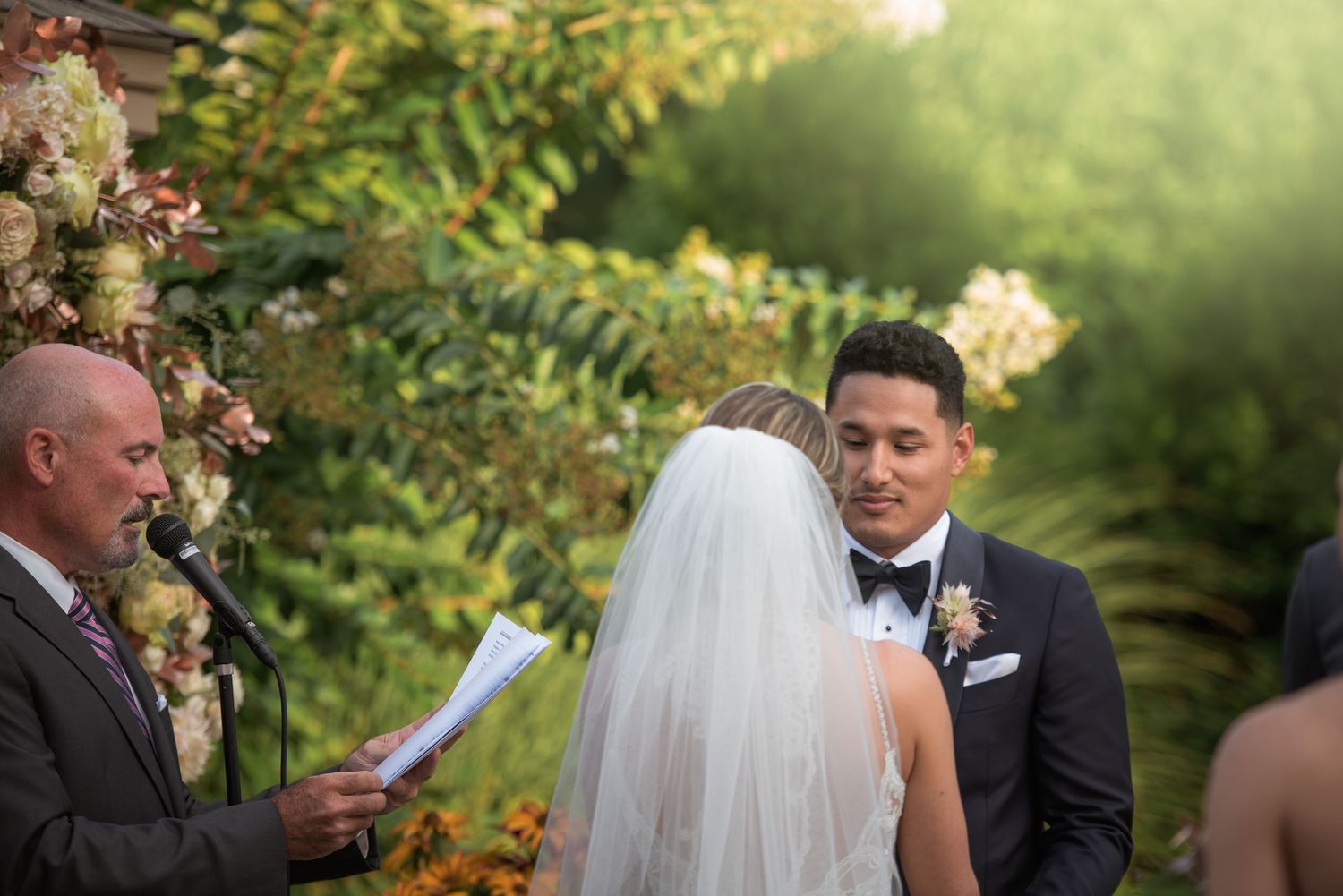 Emotional groom in navy tuxedo gazing at bride during outdoor wedding ceremony beneath lush floral arch backdrop.