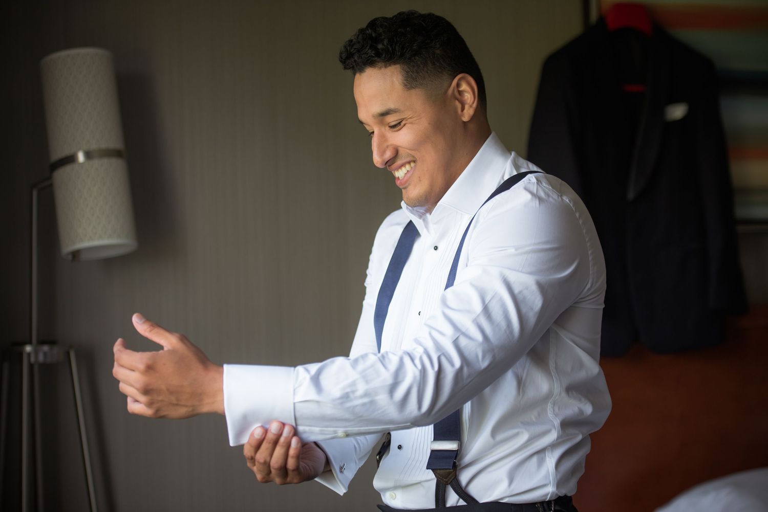 Groom in white dress shirt and suspenders smiling while fastening cufflinks during wedding day preparations in hotel room.
