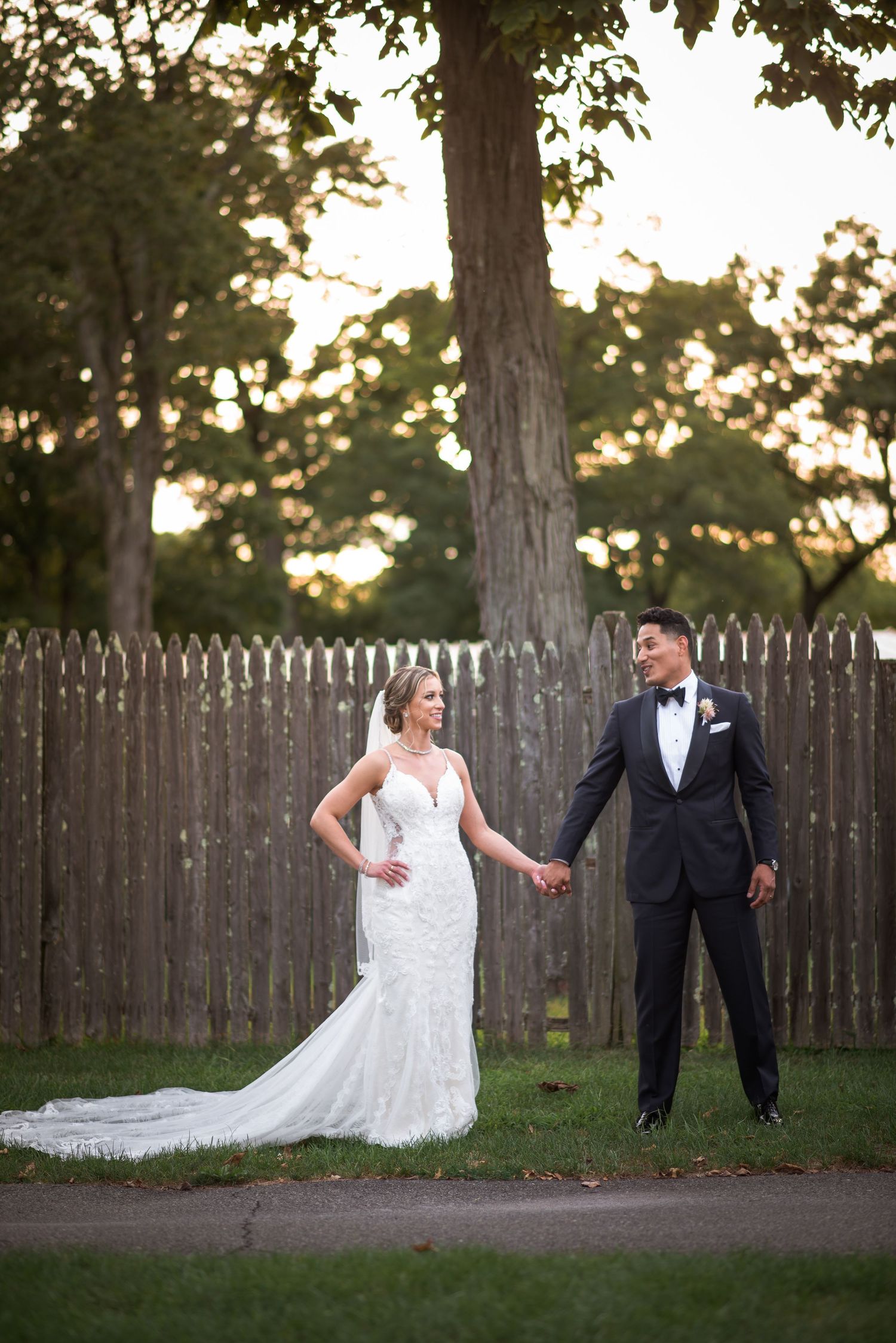Bride and groom hold hands in elegant wedding attire at golden hour outdoors.