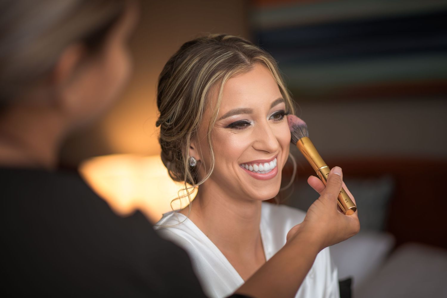 Smiling bride gets makeup applied with a brush during wedding day preparations indoors.