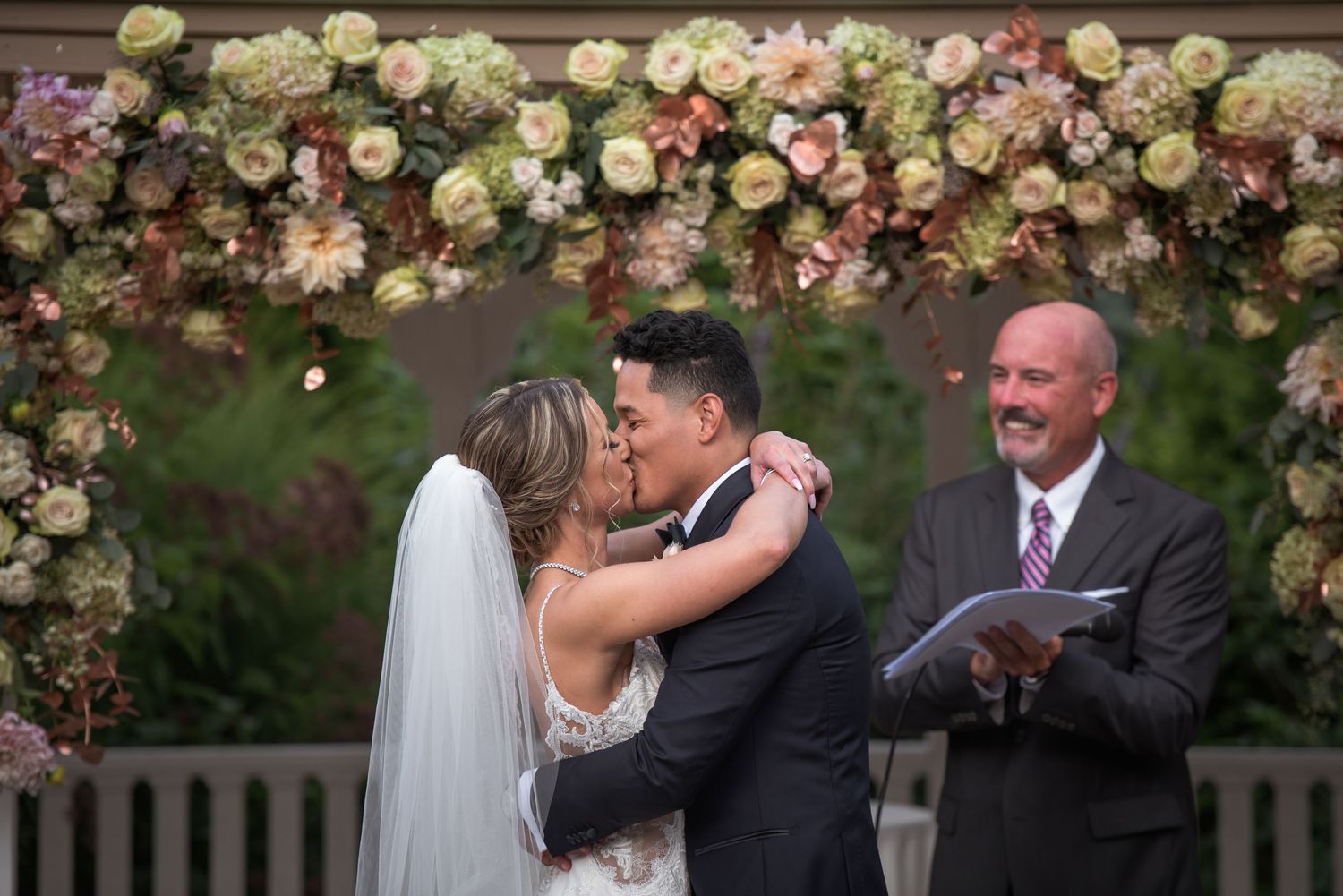 Bride and groom share first kiss under floral arch at outdoor wedding ceremony.